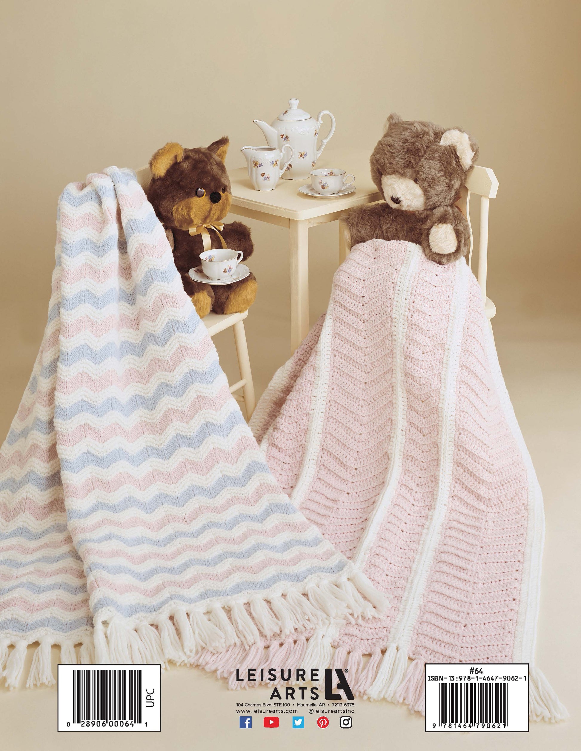 Two teddy bears sit at a small table with a toy tea set, each cozy in pastel blankets made from patterns in "Baby Afghans to Knit and Crochet" by Leisure Arts, against a light beige background.