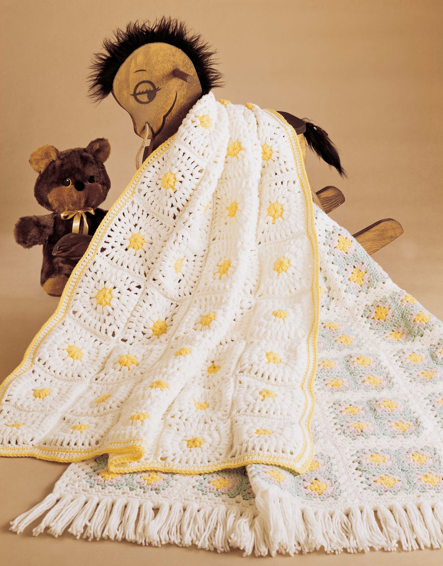 A cozy daisy-themed crocheted blanket, made from Leisure Arts' "Baby Afghans to Knit and Crochet," is draped over a wooden rocking horse beside a plush teddy bear, set against a tan background with another patterned blanket below.