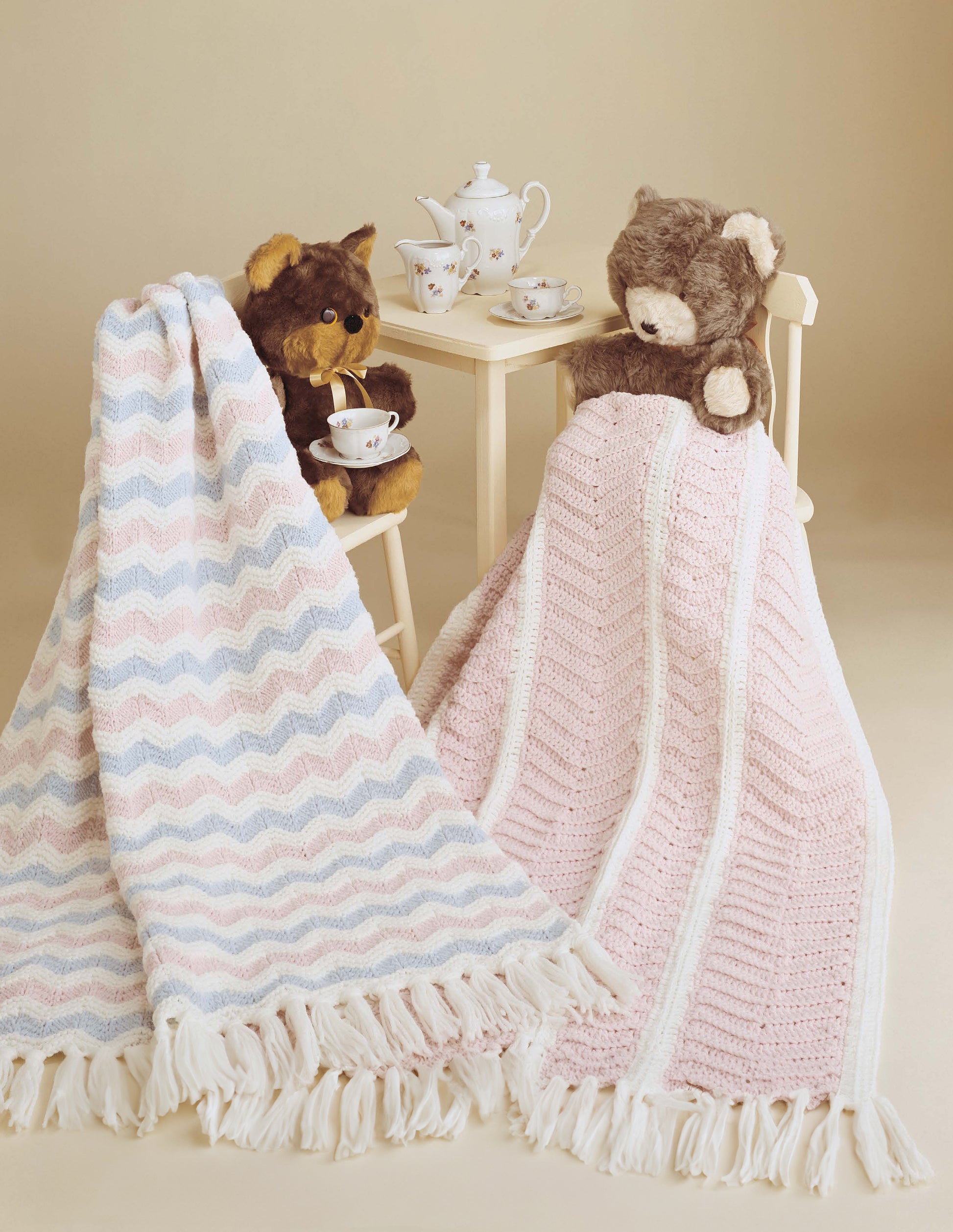 Two teddy bears enjoy a tea party at a small table, each wrapped in cozy afghans made using patterns from Leisure Arts' "Baby Afghans to Knit and Crochet," including pastel chevrons and pink-and-white granny squares.