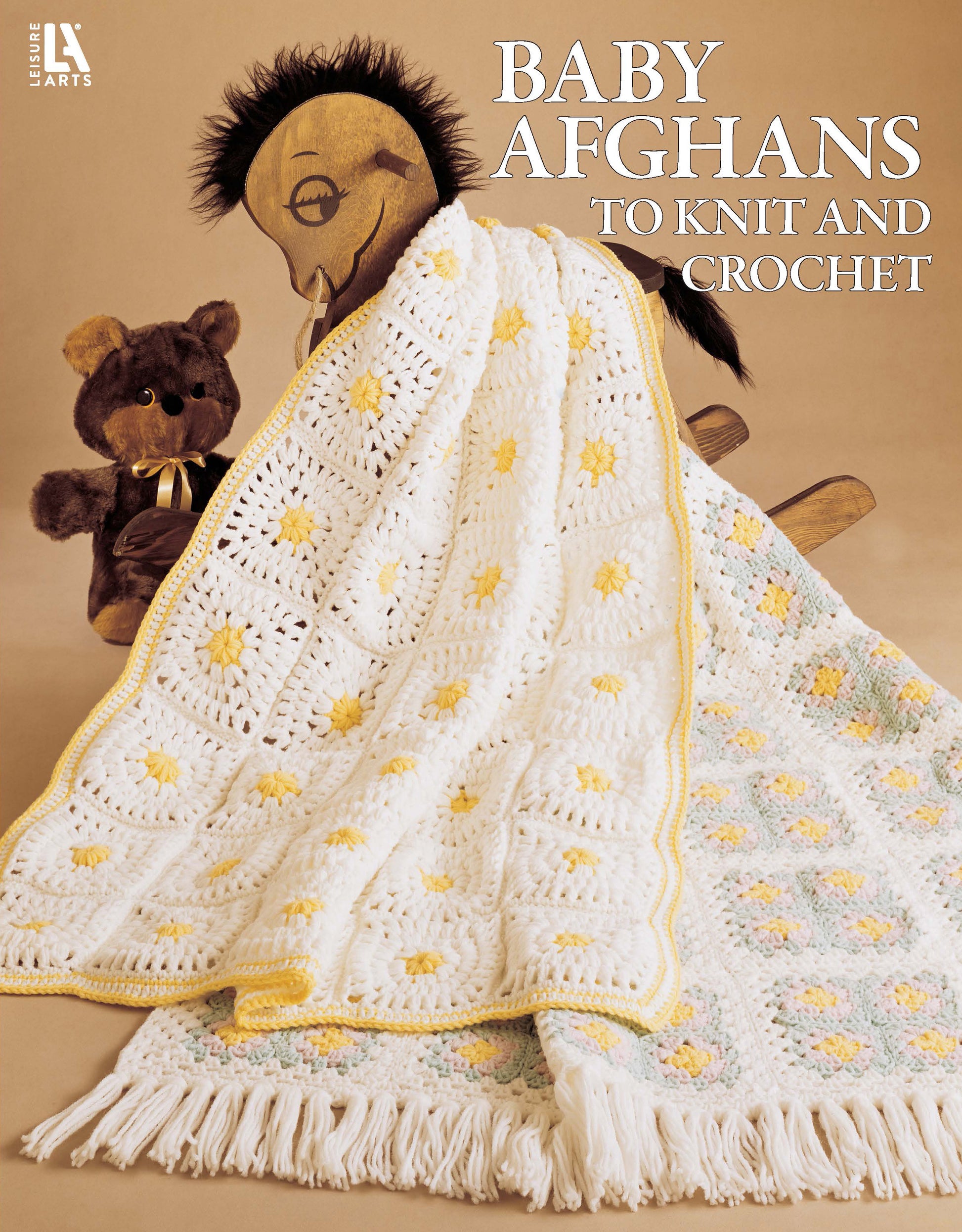 A crocheted white baby afghan with yellow and green flower accents—crafted in classic granny squares with worsted yarn—drapes over a wooden rocking horse beside a brown teddy bear. Text: Baby Afghans to Knit and Crochet by Leisure Arts.