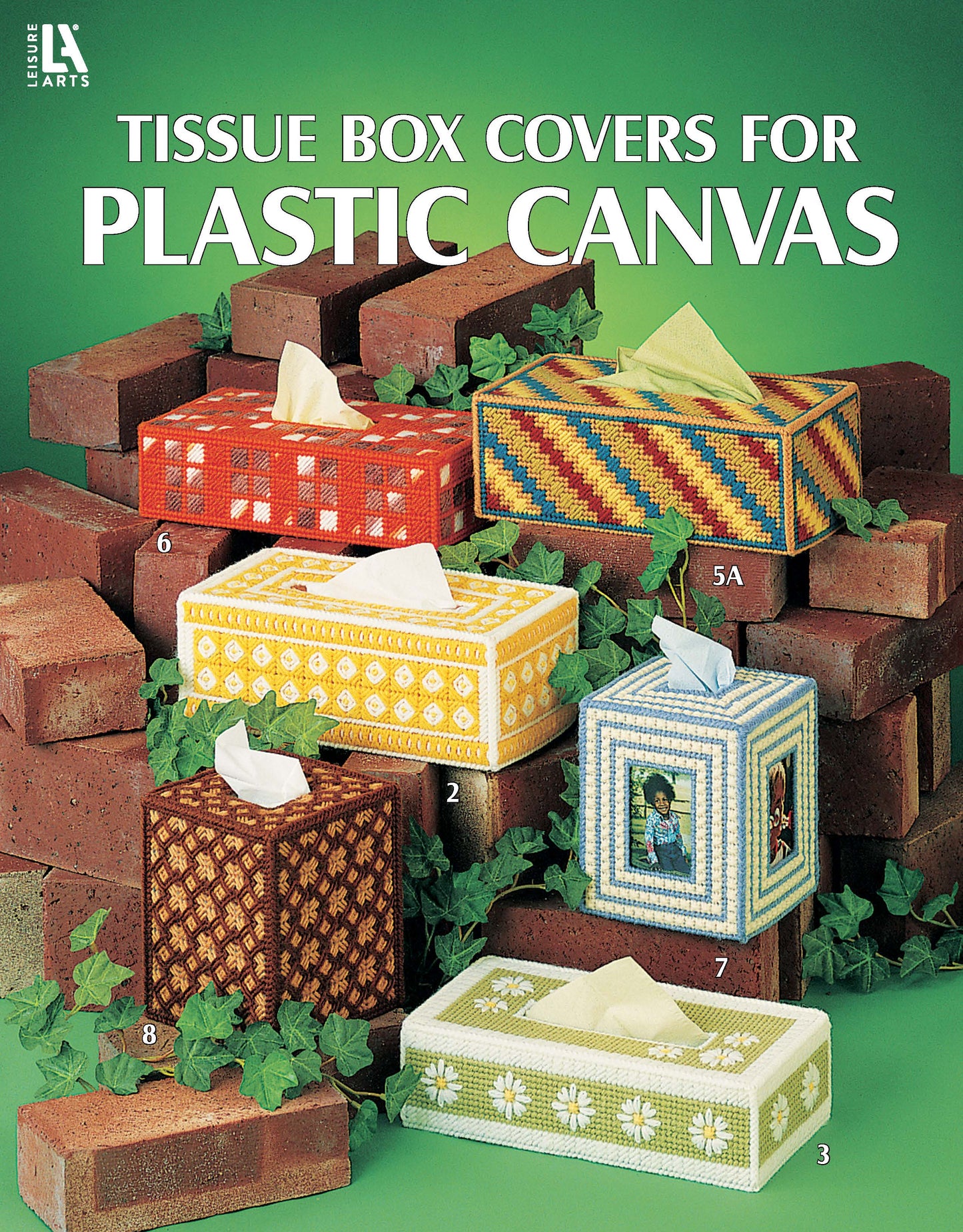 Six unique tissue box covers made with plastic canvas from Leisure Arts are shown among red bricks and green ivy. Each features decorative needlework patterns. Text: "Tissue Box Covers for Plastic Canvas.