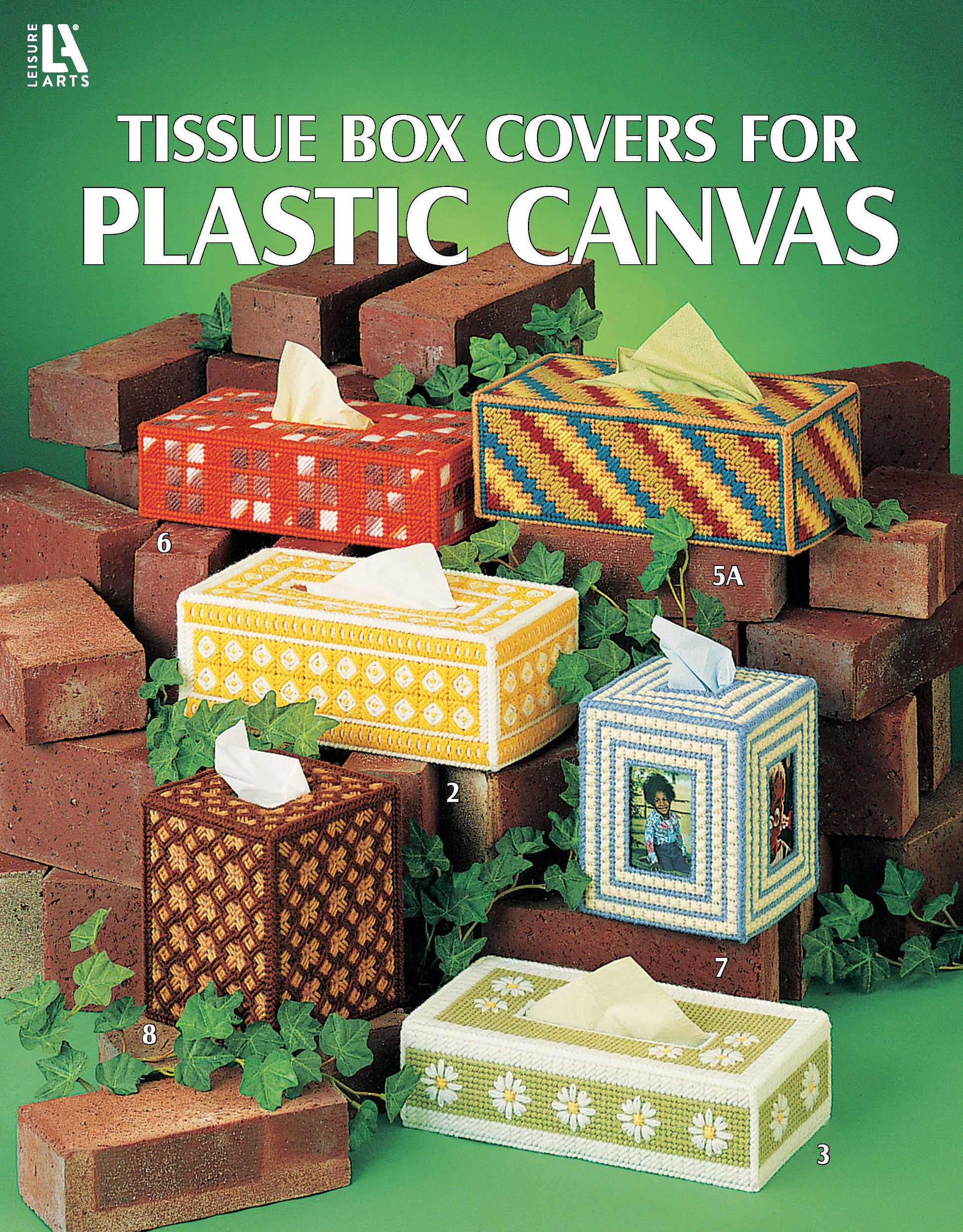 Six unique tissue box covers made with plastic canvas from Leisure Arts are shown among red bricks and green ivy. Each features decorative needlework patterns. Text: "Tissue Box Covers for Plastic Canvas.