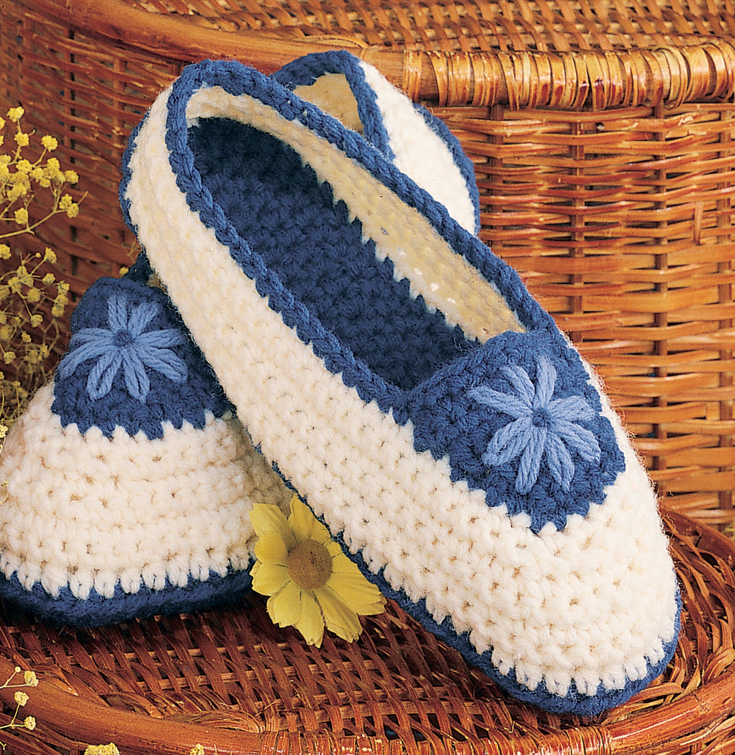 Leisure Arts Crocheted Slippers for Women & Children in blue and white, featuring blue floral accents, displayed on a wicker basket with yellow flowers.