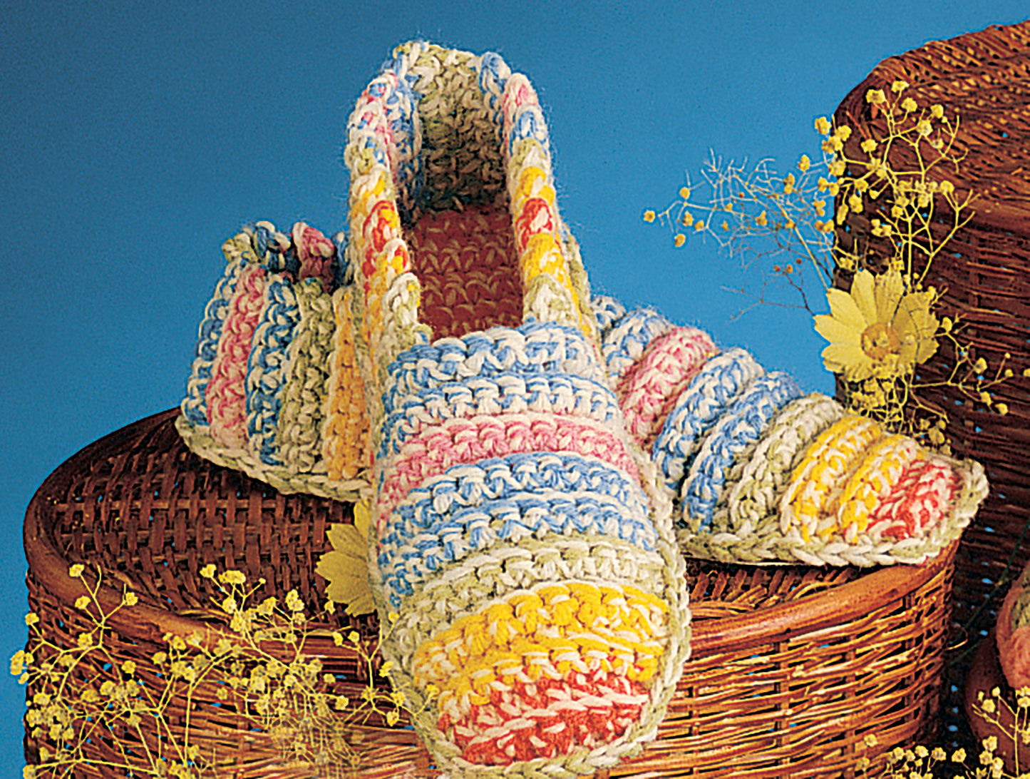 Leisure Arts Crocheted Slippers for Women & Children, featuring thick yellow, blue, pink, and white stripes, are displayed on a woven basket with yellow flowers and a bright blue background.