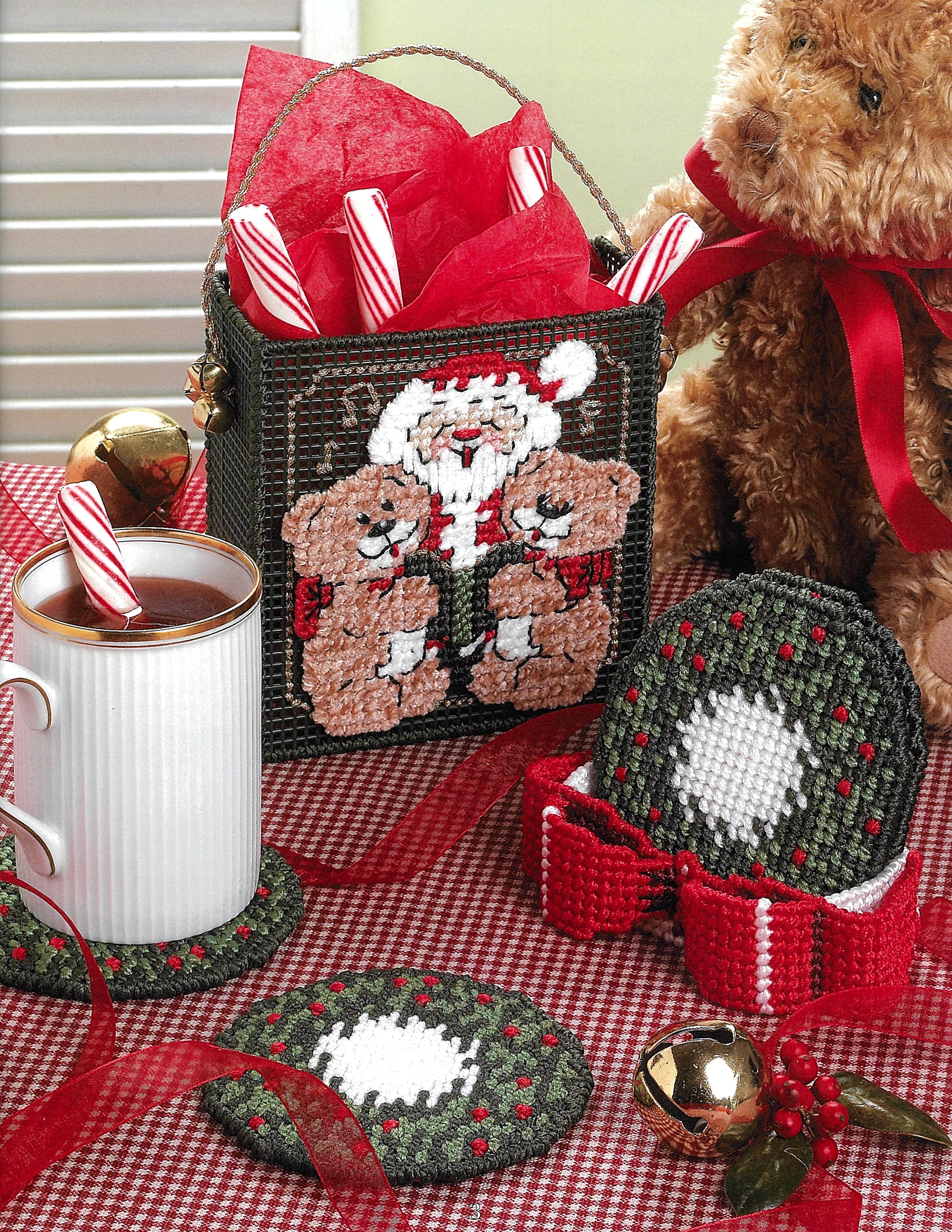 A festive table scene with a mug of cocoa, red and green woven coasters, a teddy bear, candy canes in a basket featuring Leisure Arts' "Santa, Bears, & Bells in Plastic Canvas," plus Christmas ornaments on a red checked tablecloth.