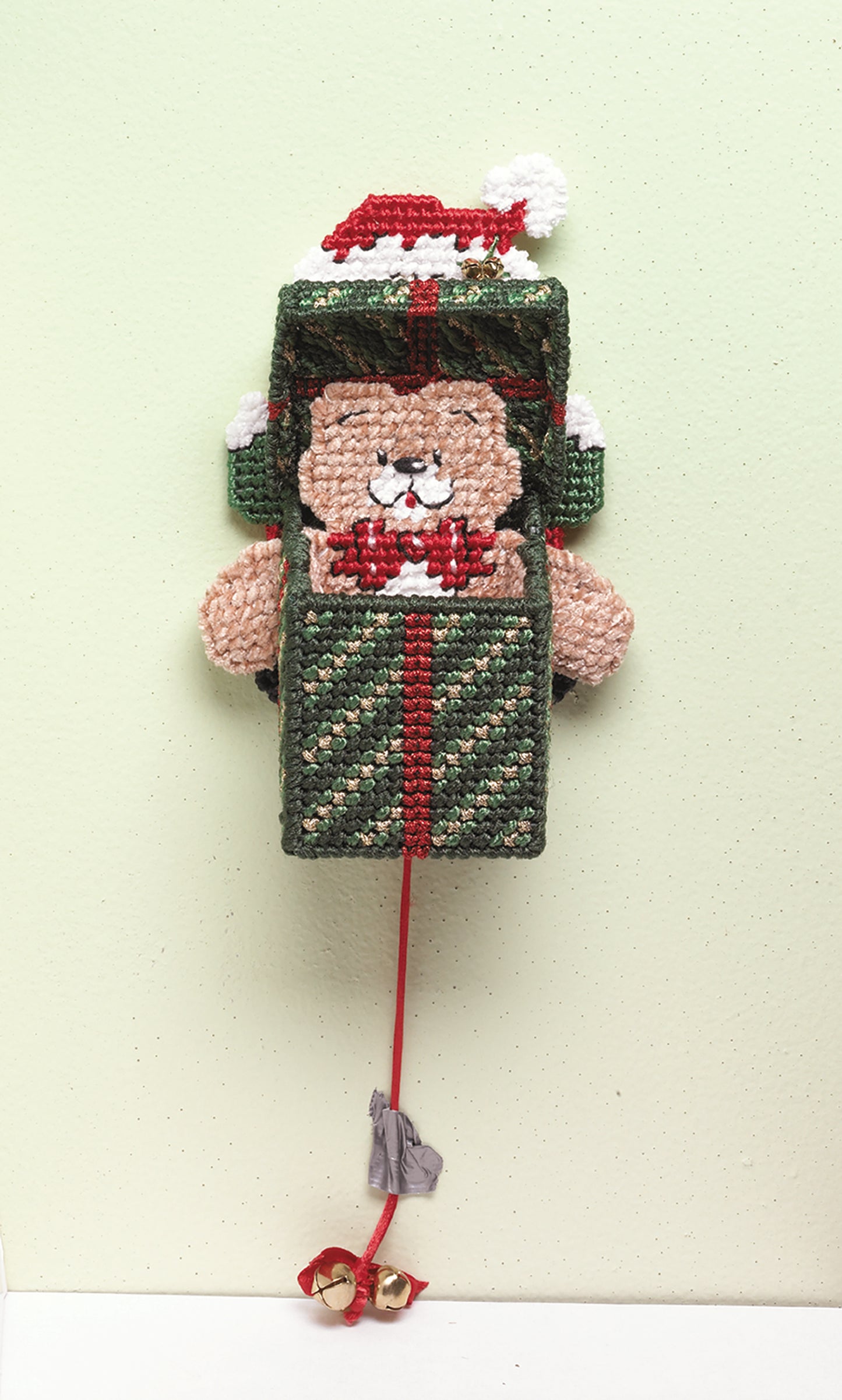 The "Santa, Bears, & Bells in Plastic Canvas" by Leisure Arts is a beginner-friendly handmade ornament featuring a bear in a Santa hat popping out of a festive gift box, complete with a red ribbon and two gold bells hanging below.