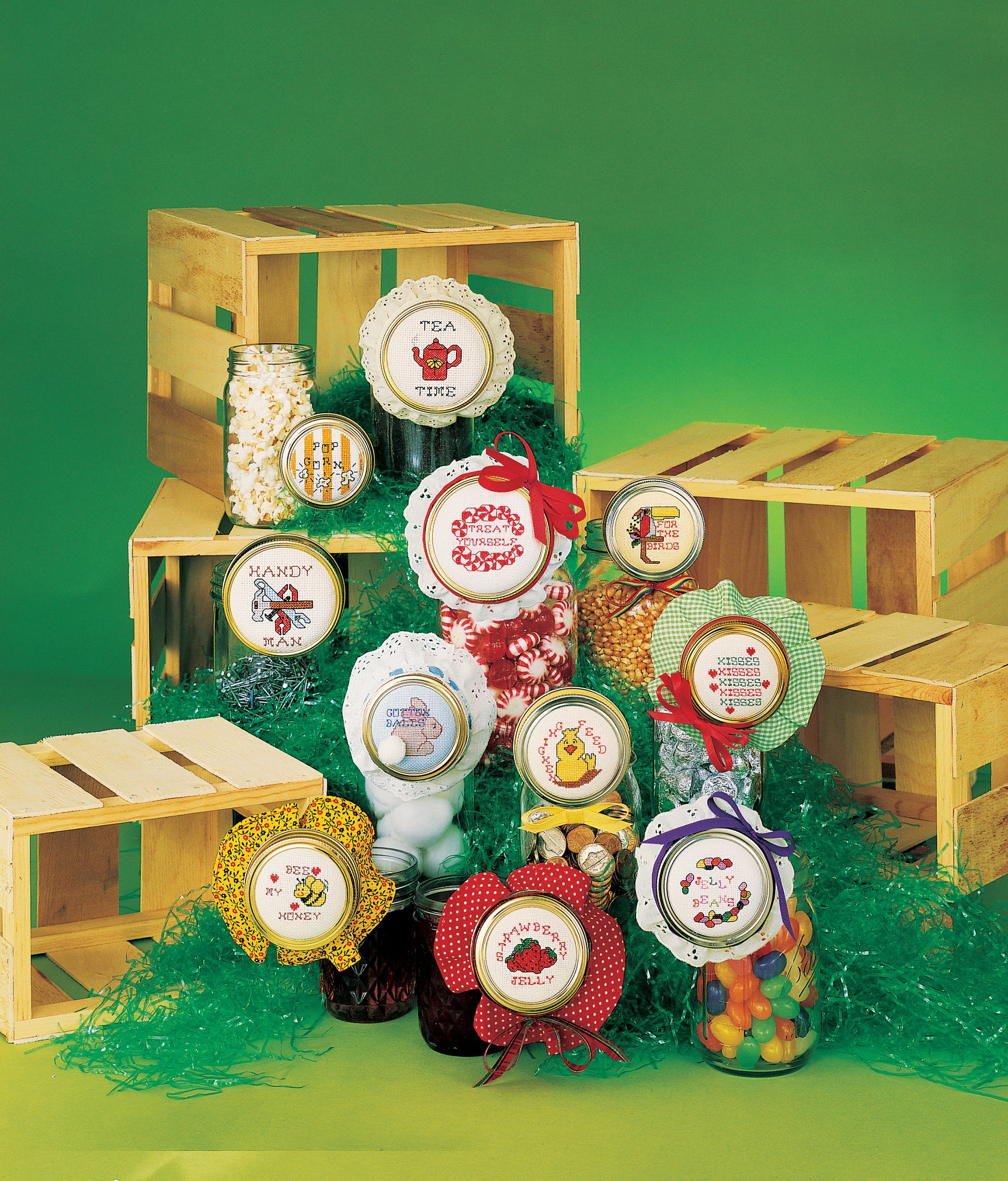 A display of jars with cross-stitched label designs from Leisure Arts’ "Jar Lids Charted for Cross Stitch Digital Download," featuring decorative fabric lids and treats, arranged on wooden crates and shredded paper against a green background.