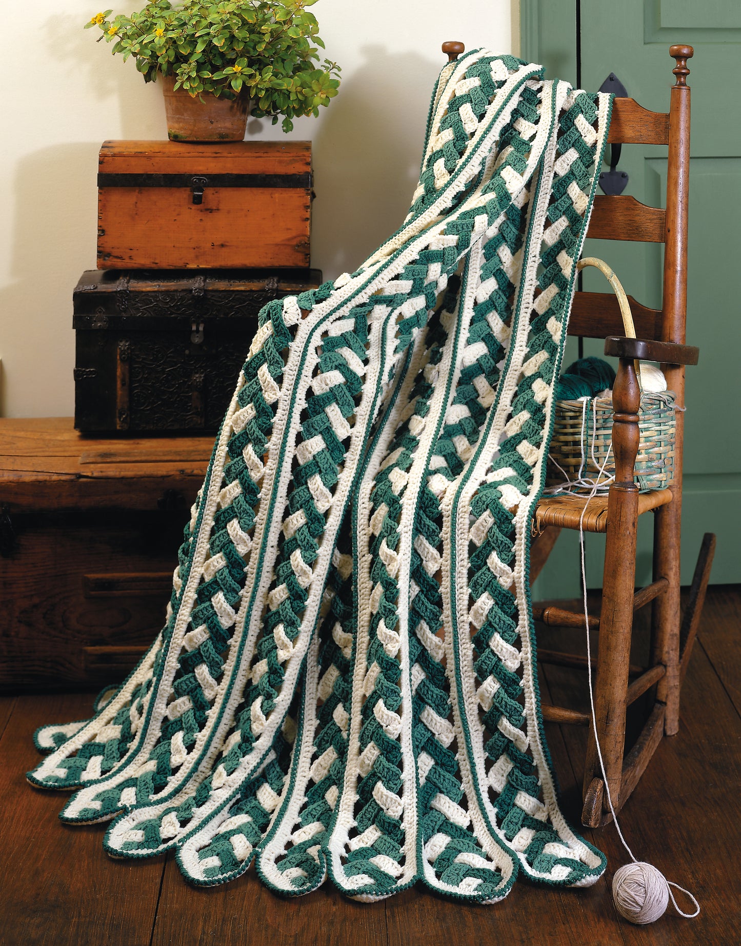 The Contest Favorite Mile-A-Minute Afghans by Leisure Arts—a green and white crocheted afghan with an interwoven pattern—drapes over a wooden chair, beside a yarn basket, with vintage trunks and a potted plant in the background.