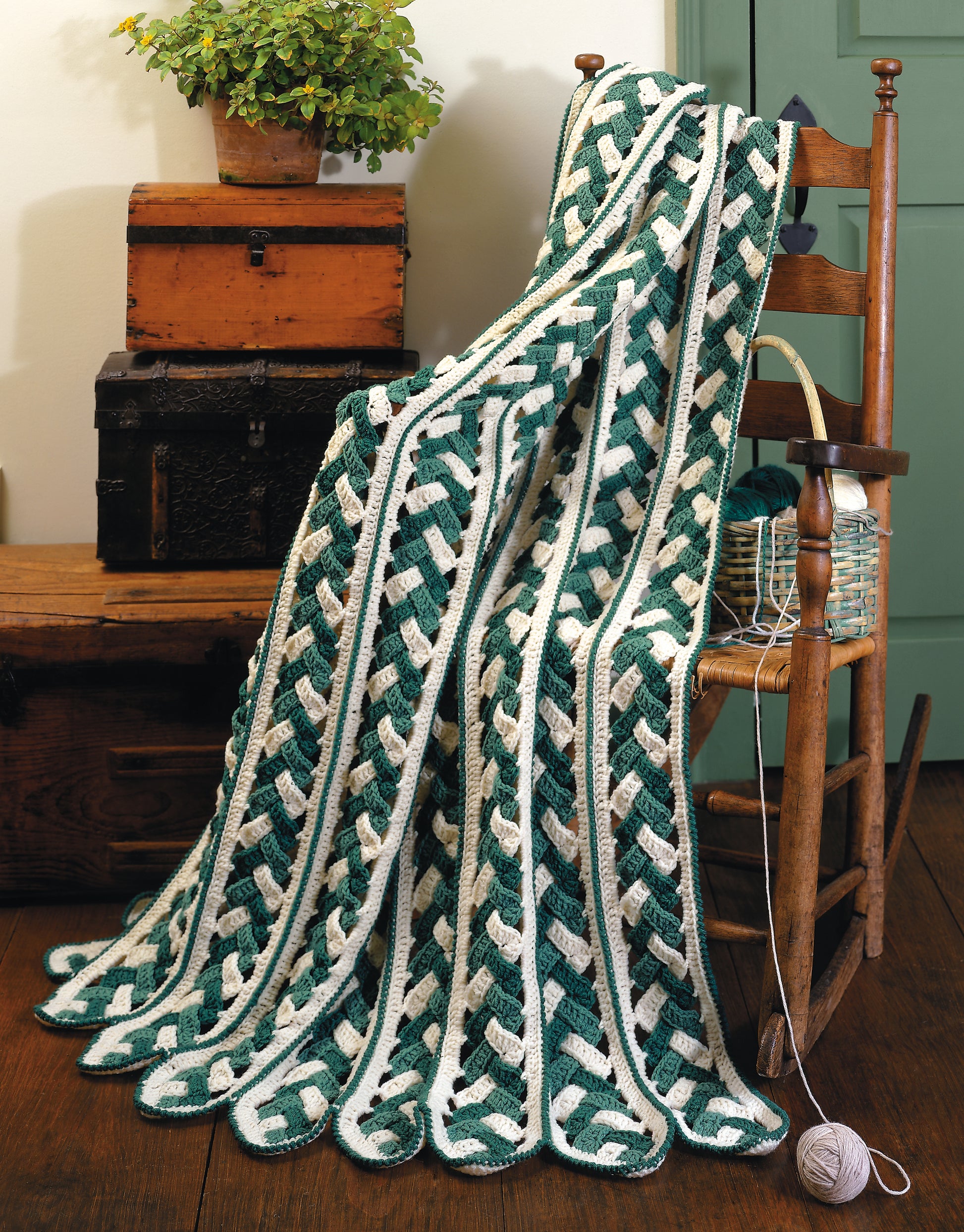 The Contest Favorite Mile-A-Minute Afghans by Leisure Arts—a green and white crocheted afghan with an interwoven pattern—drapes over a wooden chair, beside a yarn basket, with vintage trunks and a potted plant in the background.