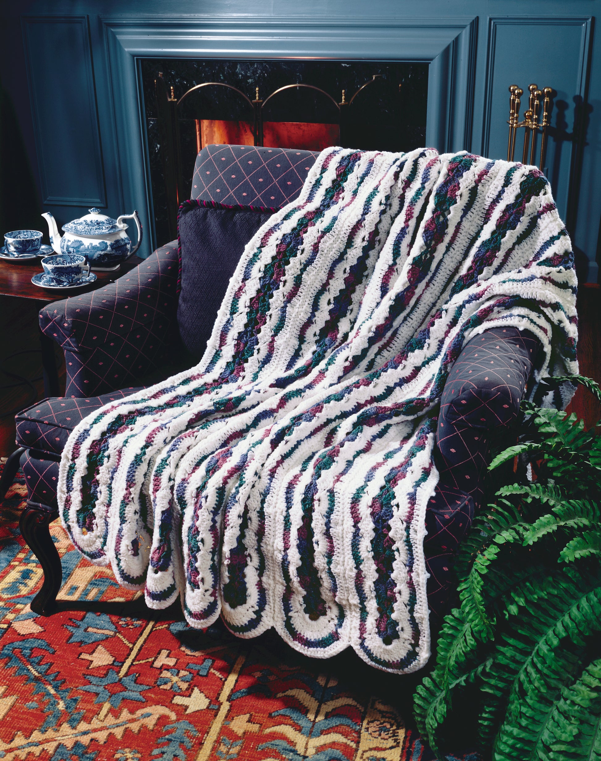 The Contest Favorite Mile-A-Minute Afghans by Leisure Arts, featuring classic white, green, and purple crochet patterns, is draped over a dark armchair next to a tea set, near a fireplace and colorful rug with a fern nearby.