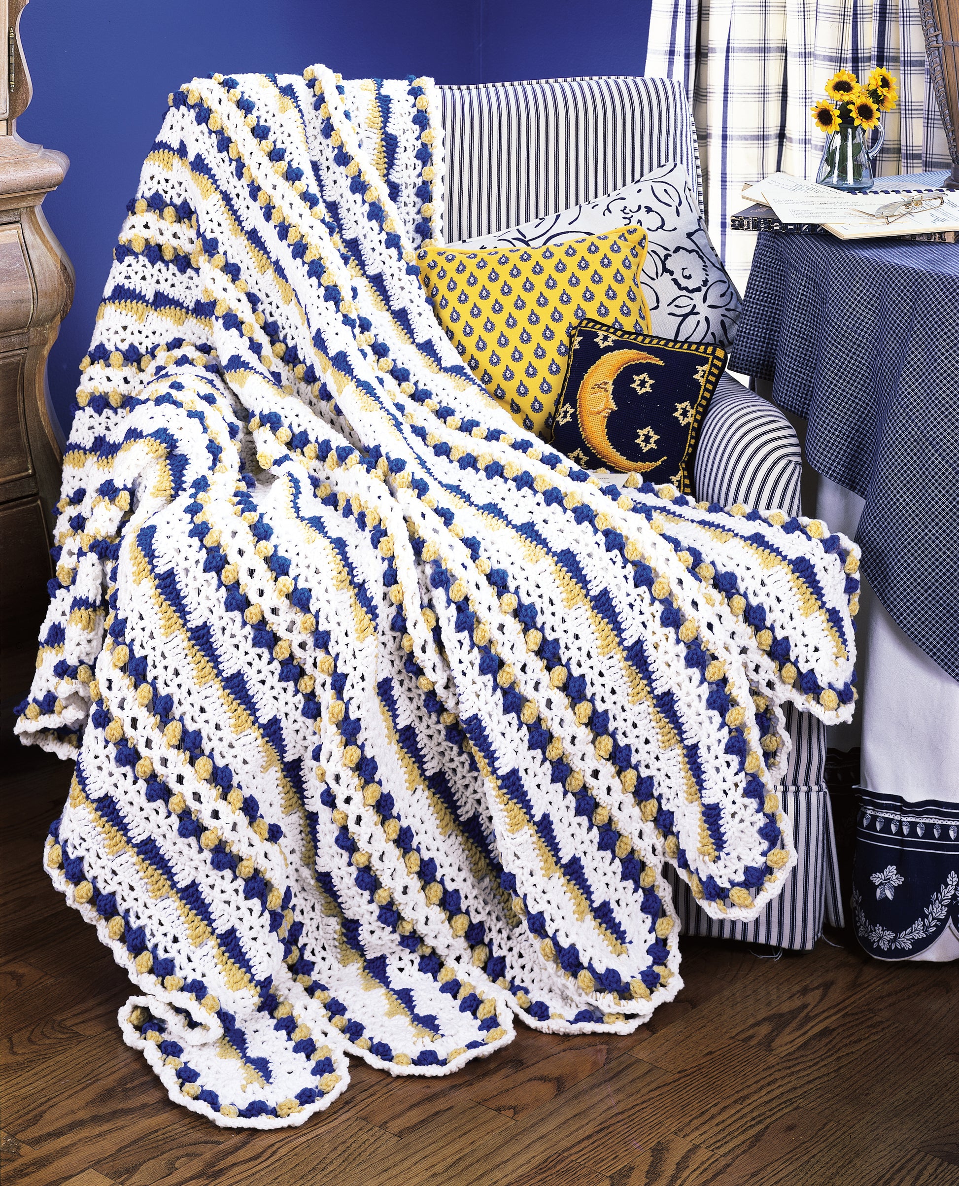 The Leisure Arts "Contest Favorite Mile-A-Minute Afghans" features a stunning crocheted afghan in white, blue, and yellow stripes draped on an armchair with matching pillows beside a table with sunflower bouquet and blue-and-white décor.