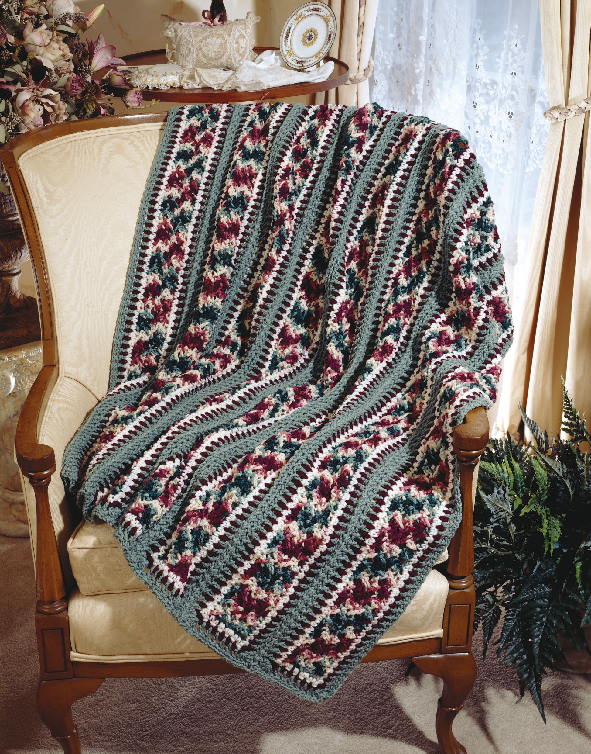 The Contest Favorite Mile-A-Minute Afghans by Leisure Arts, a patterned crocheted blanket in maroon, cream, green, and gray, is displayed on a cream-colored armchair in a cozy traditional room.