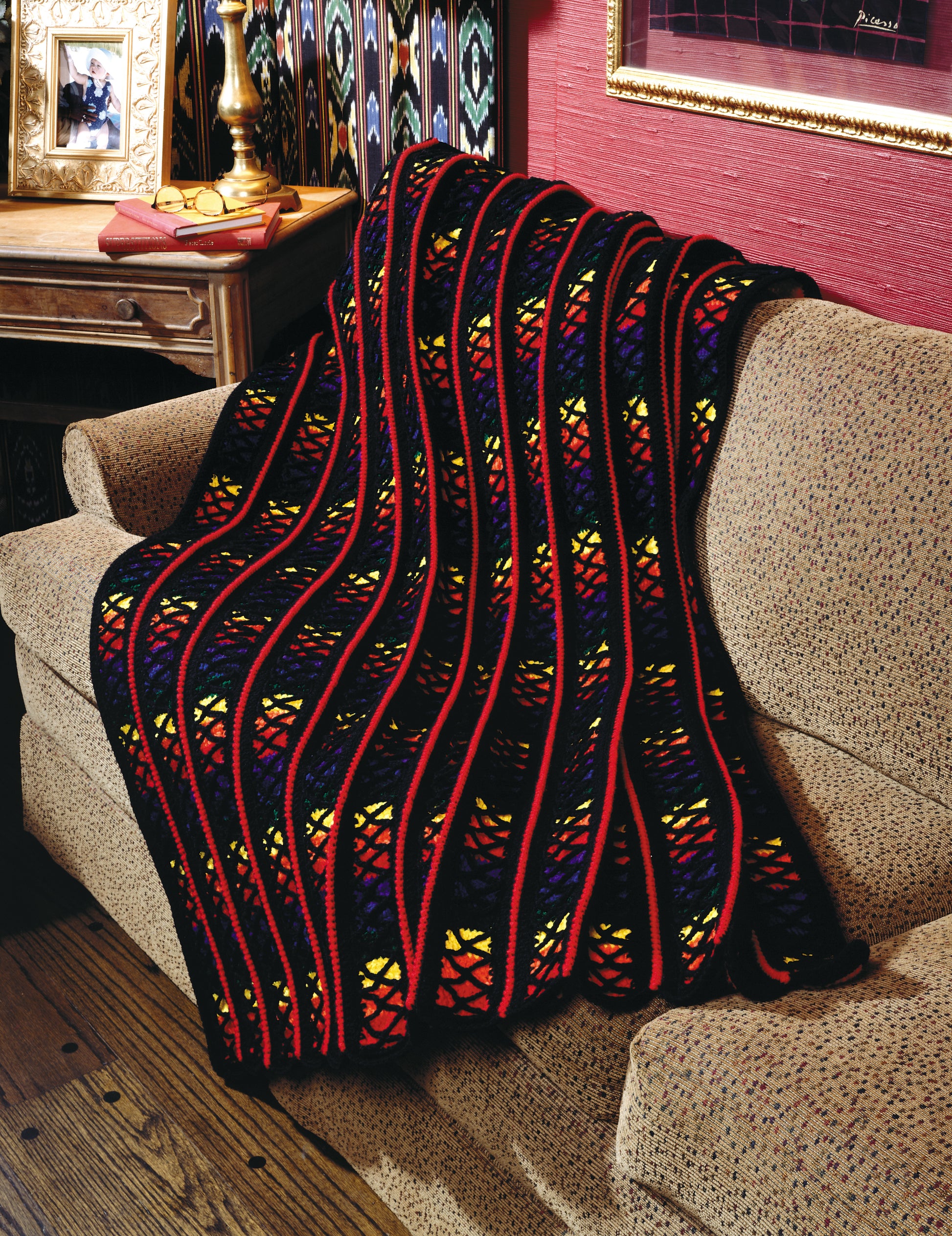 The Contest Favorite Mile-A-Minute Afghans by Leisure Arts, featuring vibrant black, red, yellow, and orange crochet designs, is draped over a beige patterned sofa in a cozy, warmly decorated living room.