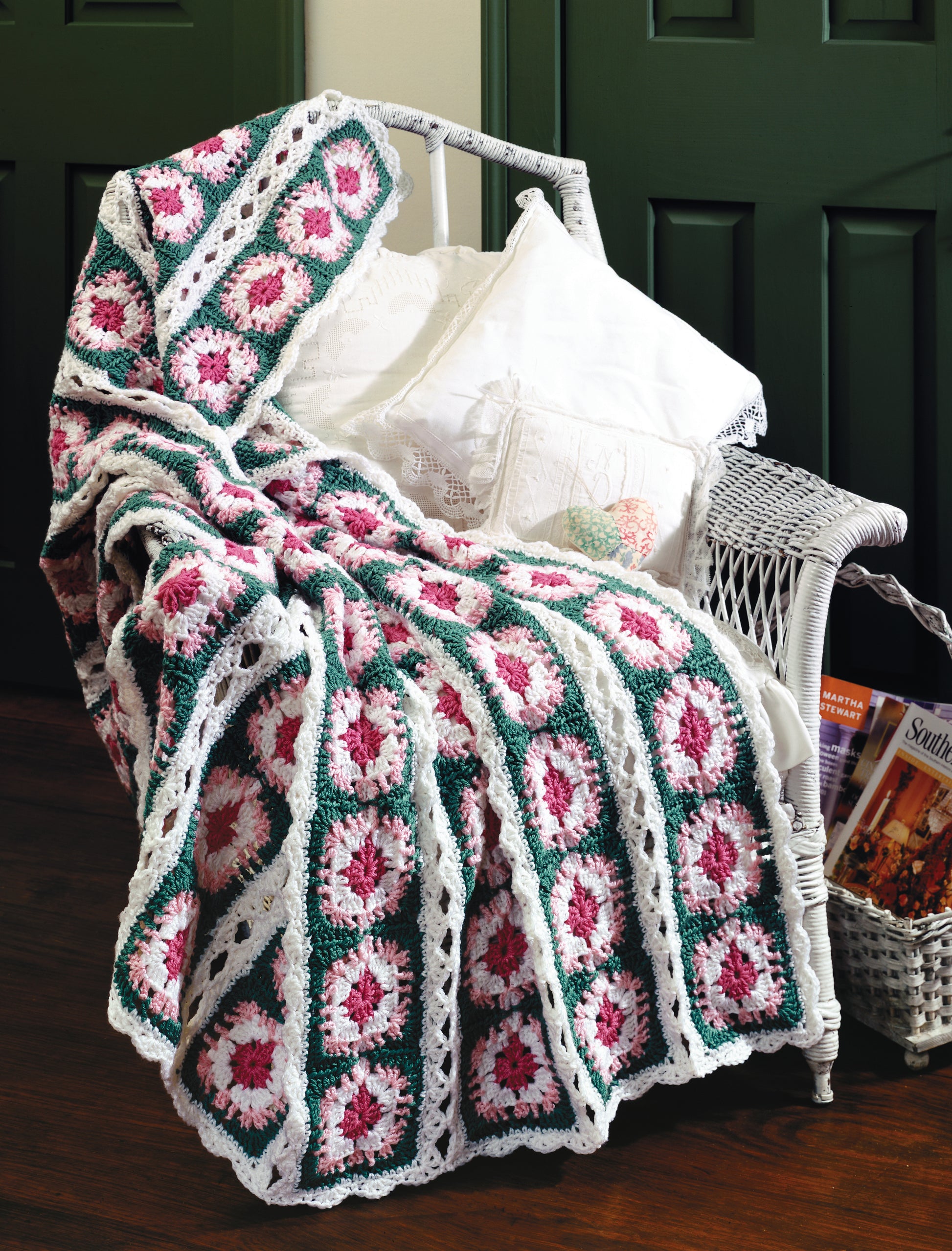 A white wicker chair holds white pillows and a crocheted blanket made with the Contest Favorite Mile-A-Minute Afghans Digital Download by Leisure Arts. A magazine rack with books is attached, and green doors are seen in the background.
