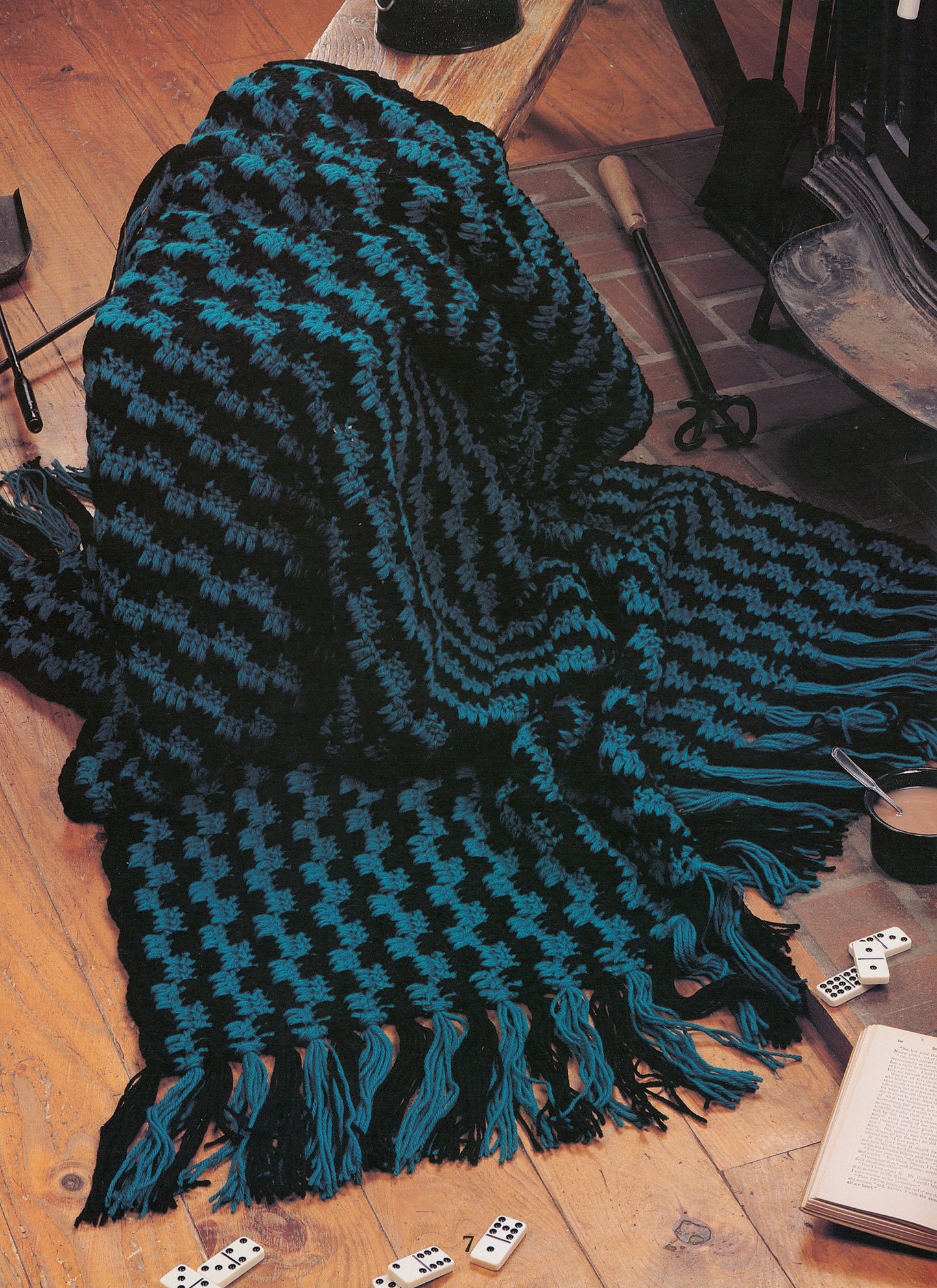 The Rustic Warmth—11 Afghans by Leisure Arts, featuring 9 crochet and 2 knit designs, is shown in blue and black houndstooth with fringe, draped on a wooden floor near a fireplace with dominoes and a book nearby.