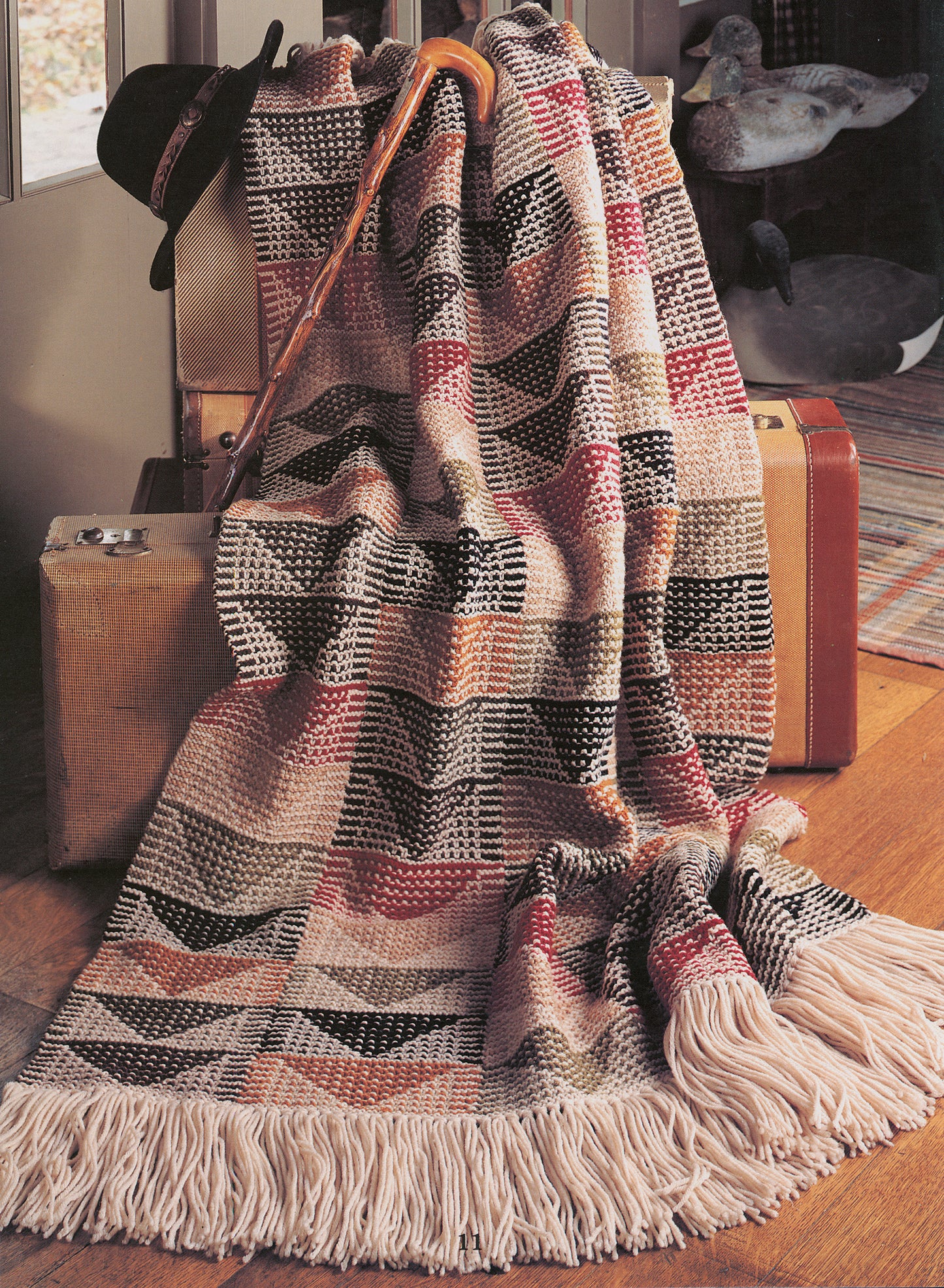 A crocheted blanket from Leisure Arts' "Rustic Warmth—11 Afghans" in earthy tones drapes over vintage suitcases. A cane, black hat, wooden duck figurines, a rug, and part of a door are also visible.