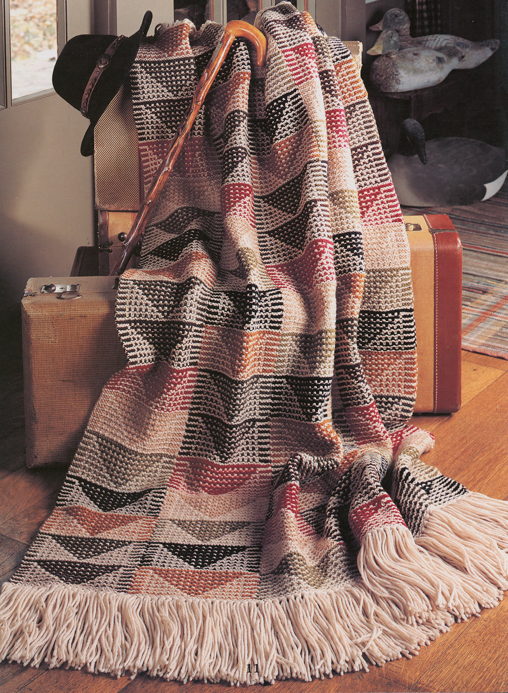 A crocheted blanket from Leisure Arts' "Rustic Warmth—11 Afghans" in earthy tones drapes over vintage suitcases. A cane, black hat, wooden duck figurines, a rug, and part of a door are also visible.