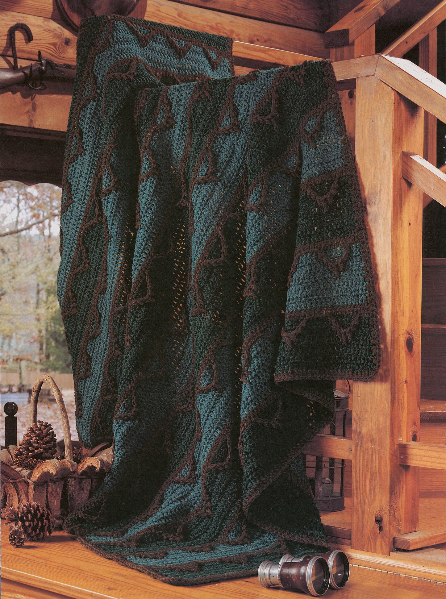A dark green and brown mountain-themed afghan from "Rustic Warmth—11 Afghans - 9 to Crochet, 2 to Knit" by Leisure Arts drapes over a wooden railing in a cabin, with pinecones and binoculars nearby.
