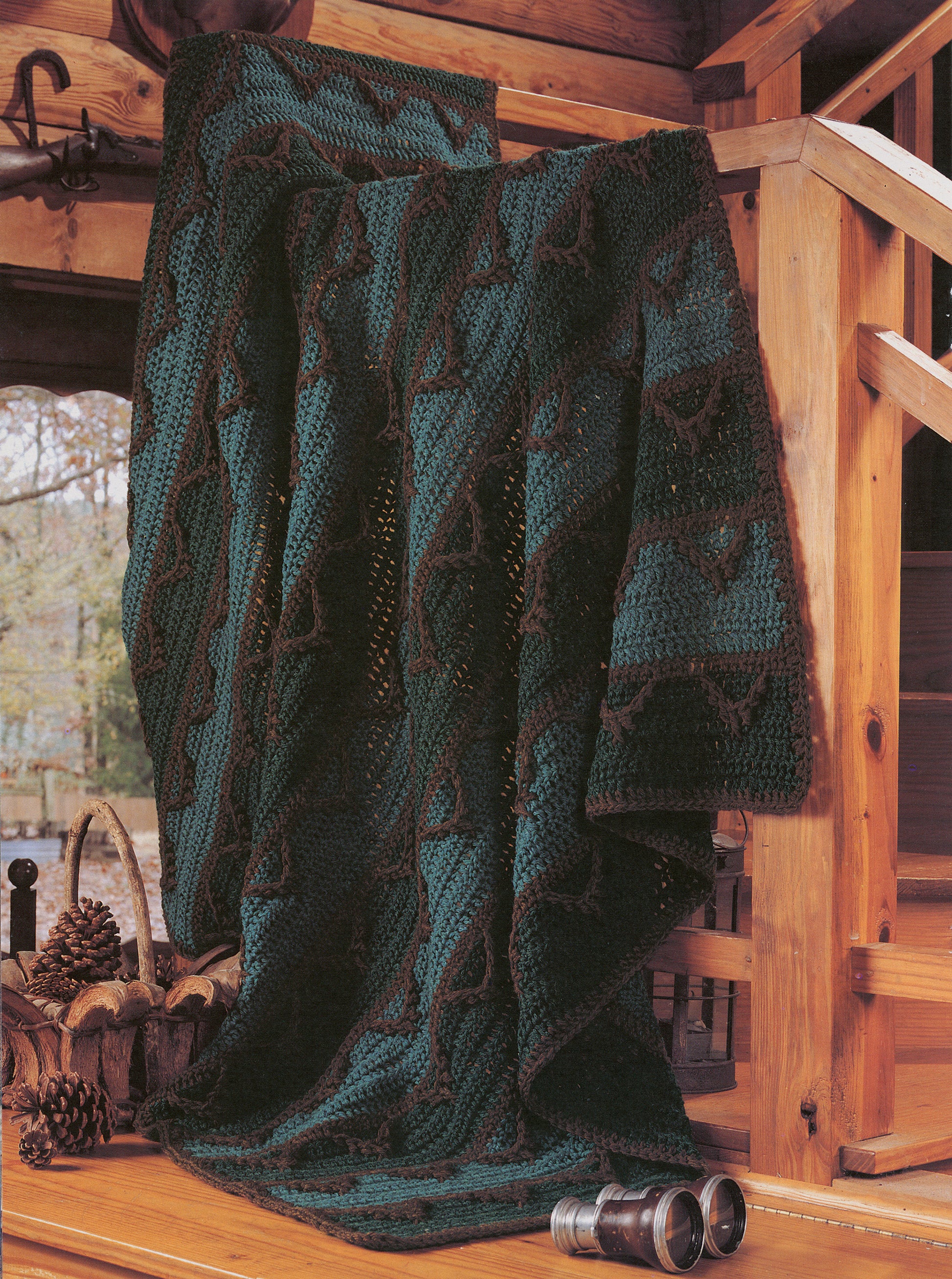 A dark green and brown mountain-themed afghan from "Rustic Warmth—11 Afghans - 9 to Crochet, 2 to Knit" by Leisure Arts drapes over a wooden railing in a cabin, with pinecones and binoculars nearby.