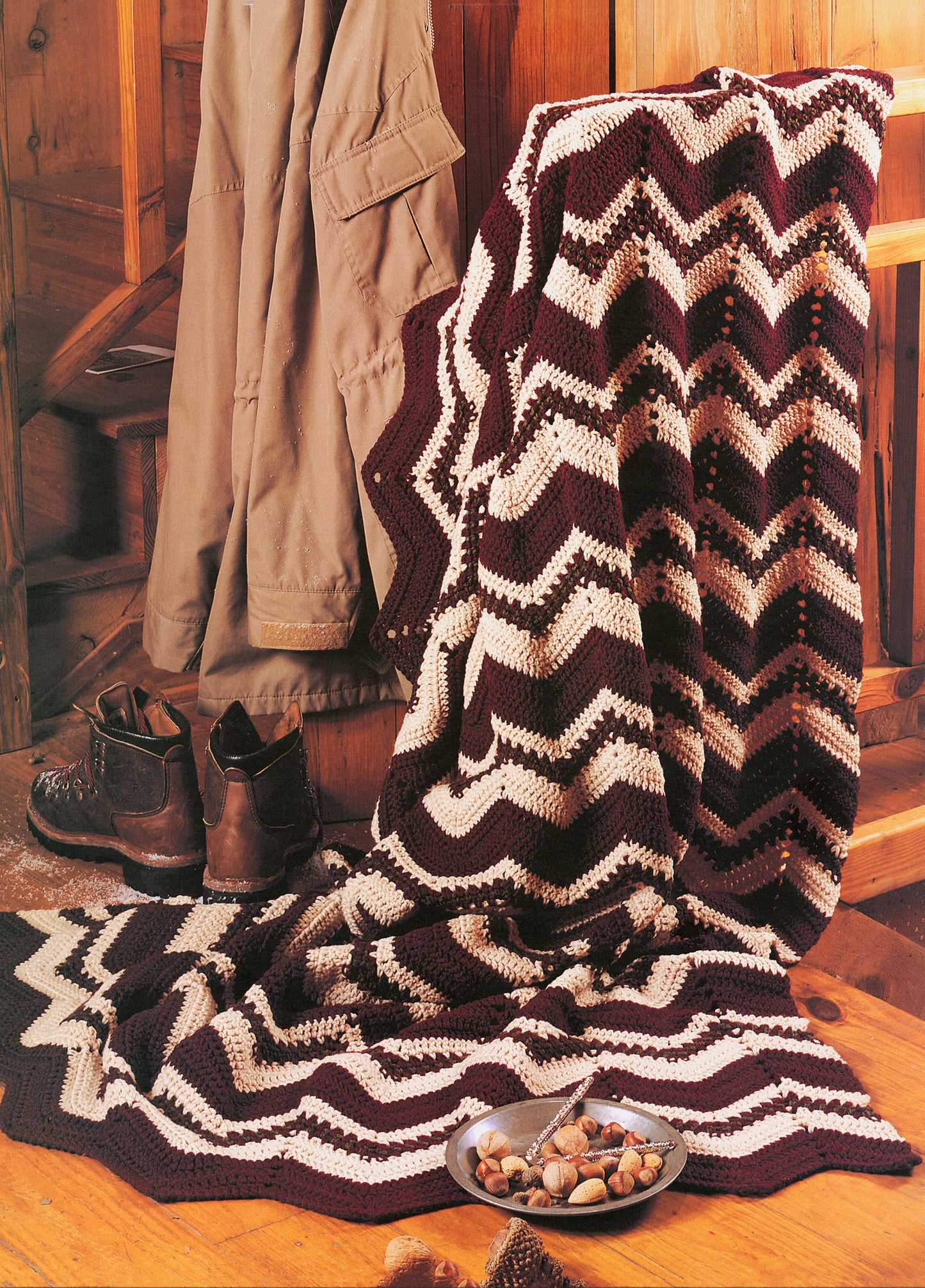 A striped, zigzag-patterned afghan from Leisure Arts’ Rustic Warmth—11 Afghans (9 to crochet, 2 to knit) drapes over a wooden chair beside outerwear and boots in a cozy cabin room, with a bowl of nuts on the table.