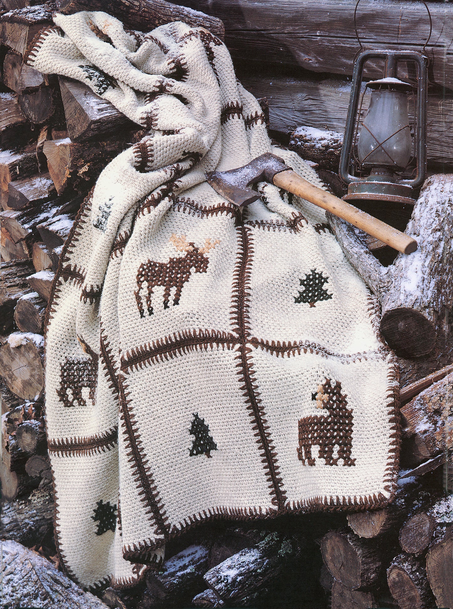 The Rustic Warmth—11 Afghans by Leisure Arts, featuring nature-inspired crochet designs like moose and trees, is draped over stacked firewood with a lantern and hatchet nearby.