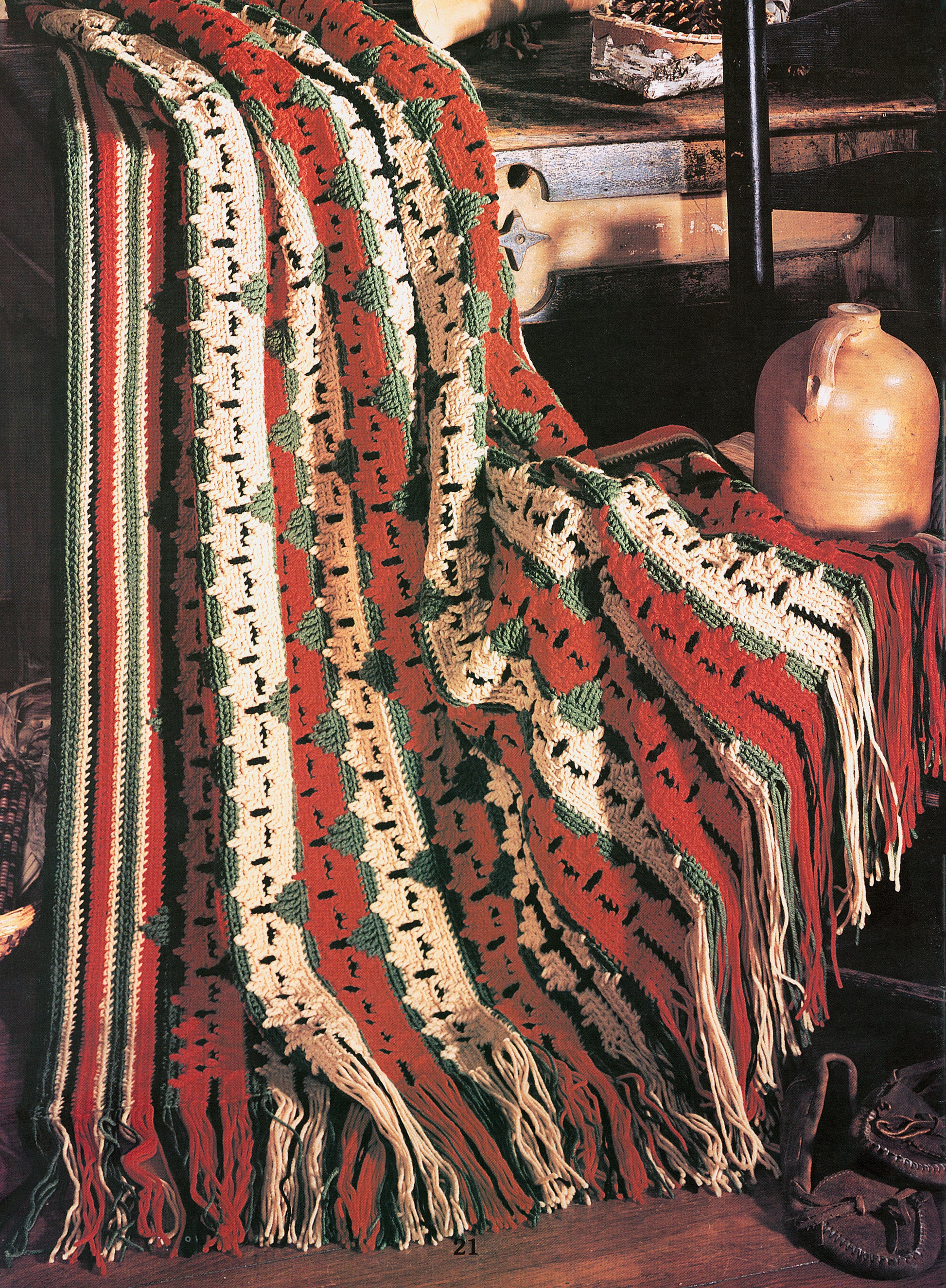 A crocheted afghan from Leisure Arts’ “Rustic Warmth—11 Afghans” features red, white, and green stripes with fringe and is draped over a wooden chair beside a pottery jug and brown shoes in a cozy, rustic setting.