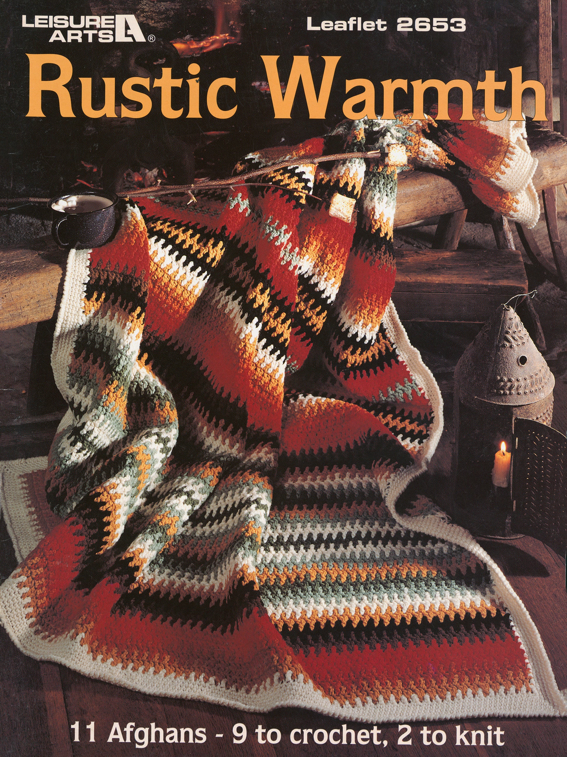 A cozy afghan with zigzag patterns in autumn hues is draped over a wooden chair beside a lantern. Text: “Rustic Warmth—11 Afghans: 9 crochet, 2 knit, by Leisure Arts.”.