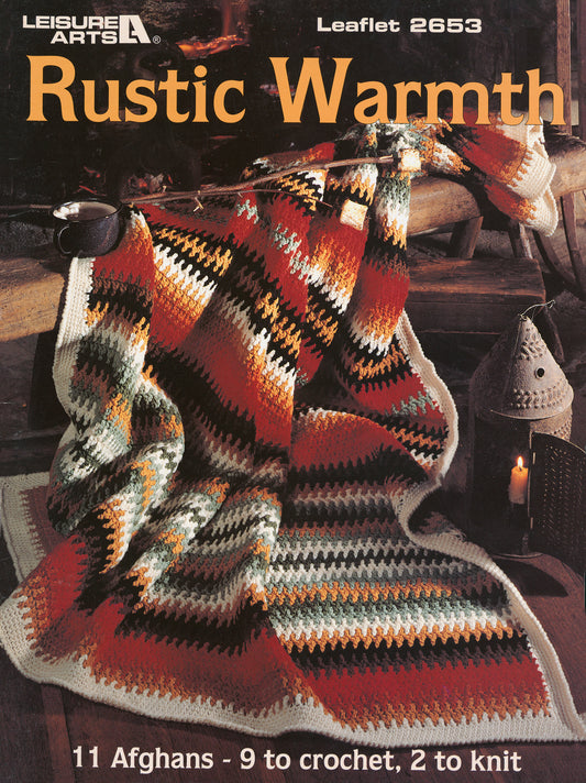 A cozy afghan with zigzag patterns in autumn hues is draped over a wooden chair beside a lantern. Text: “Rustic Warmth—11 Afghans: 9 crochet, 2 knit, by Leisure Arts.”.
