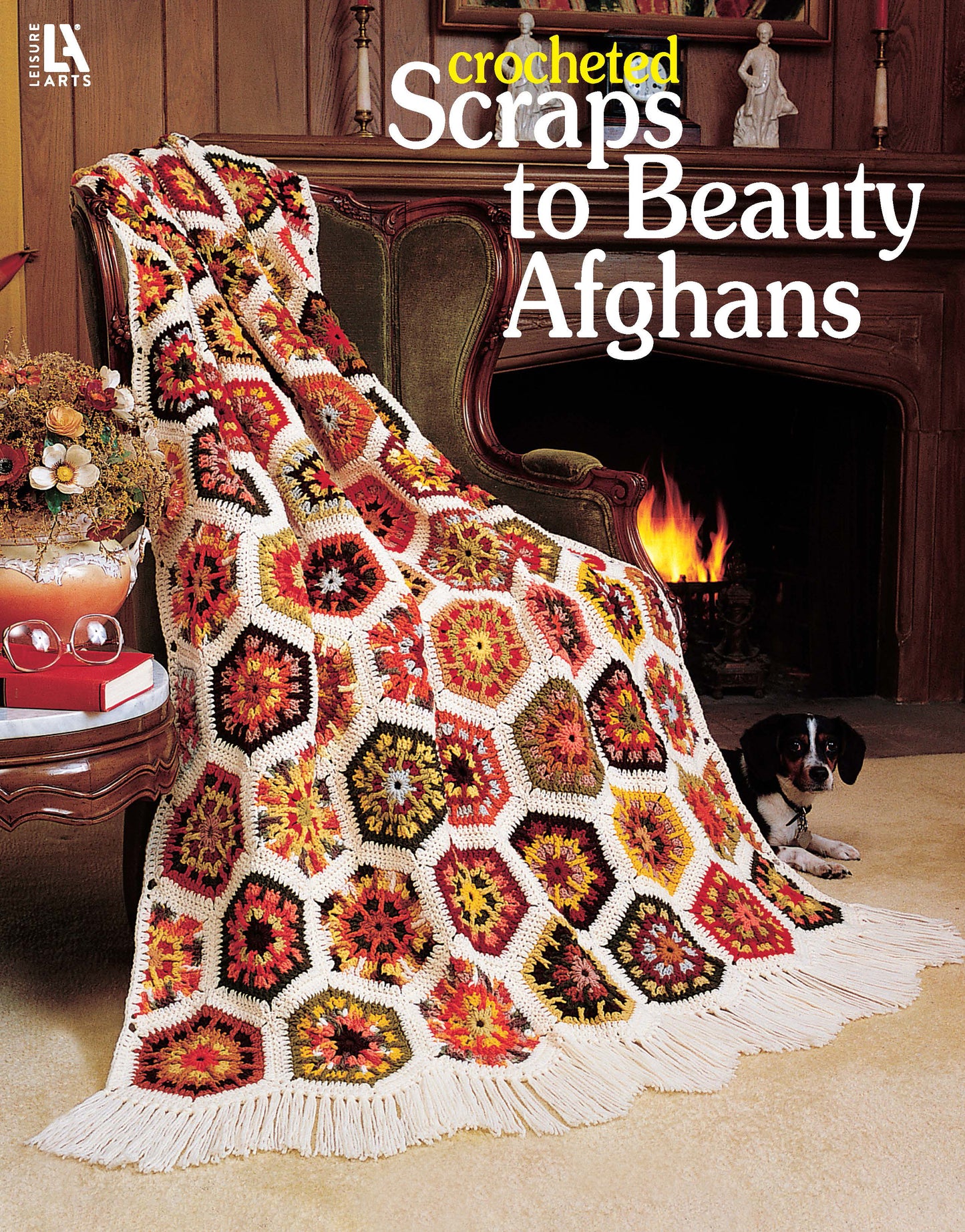 A vibrant afghan from leftover yarn, featuring colorful hexagons and fringe, drapes over a chair by the fire while a black and white dog rests nearby. Text: “Leisure Arts Crocheted Scraps to Beauty Afghans Digital Download.”.