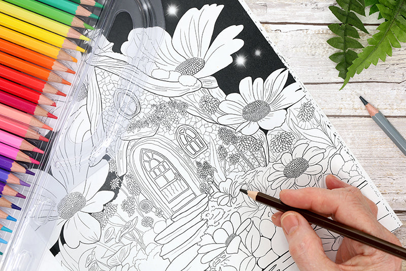 A hand colors a detailed page from Leisure Arts’ “Enchanted Gardens—36 Magical Fantasy Designs for Adult Coloring” with colored pencils. An open box of assorted pencils rests beside it, making this set ideal for beginners and relaxation.