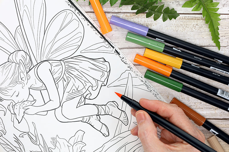 A hand colors a fairy illustration from Leisure Arts' Enchanted Gardens—36 Magical Fantasy Designs for Adult Coloring with an orange marker, surrounded by colored markers and green fern leaves on a wooden table.