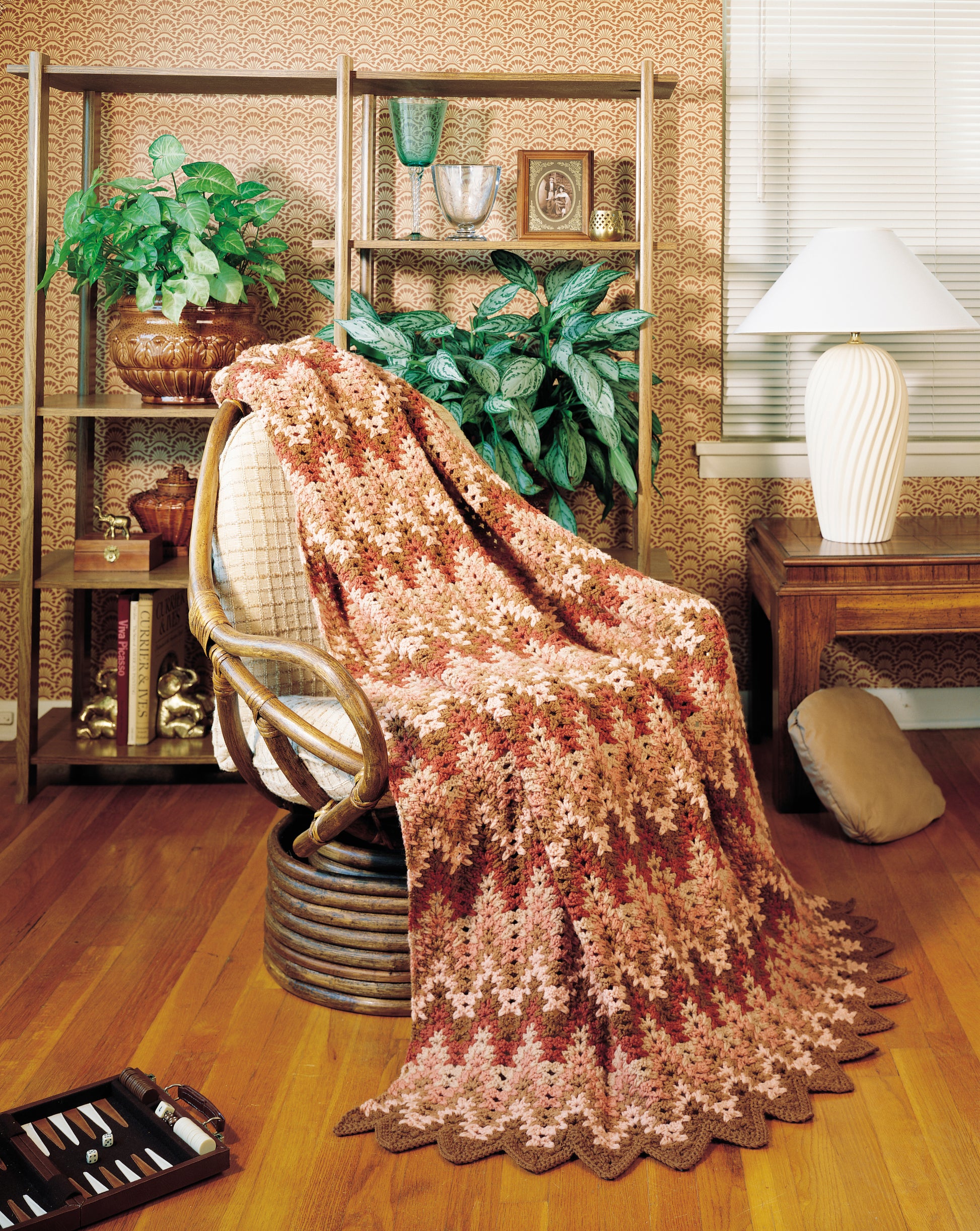 The Afghan Parade by Leisure Arts, a crocheted blanket in brown and pink worsted weight yarn, decorates a cozy chair on a wooden floor amid houseplants, decorative shelves, a table lamp, and patterned wallpaper.