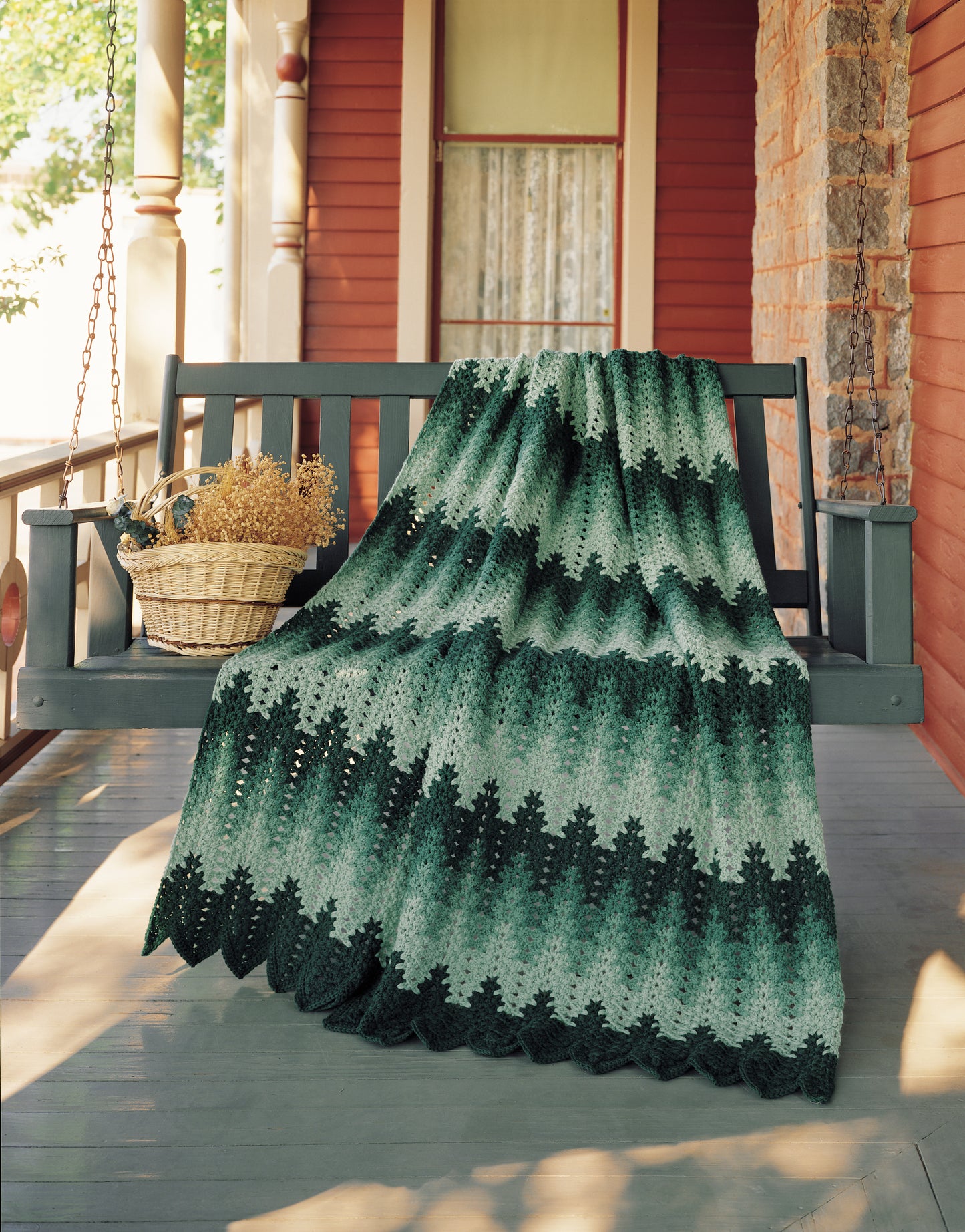 The Afghan Parade by Leisure Arts, a green and black chevron-patterned crocheted blanket made with worsted weight yarn, is draped over a gray porch swing beside a wicker basket of dried flowers on a red-and-white columned porch.