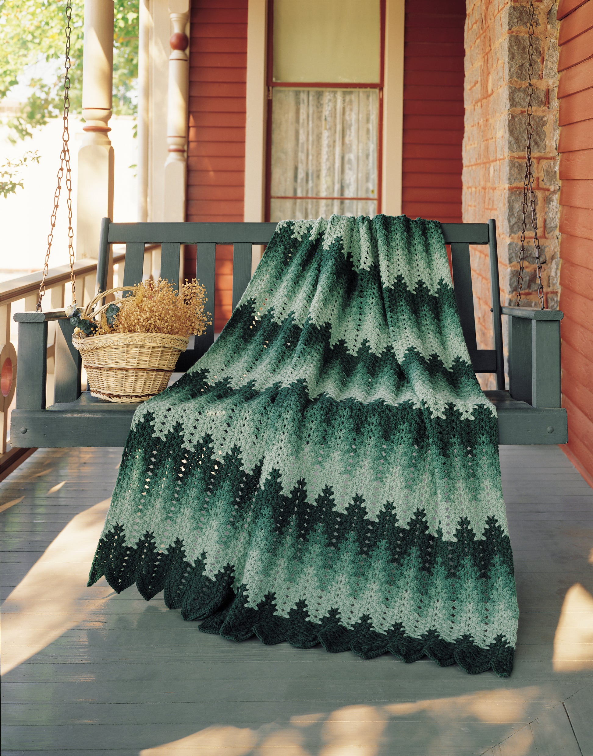 The Afghan Parade by Leisure Arts, a green and black chevron-patterned crocheted blanket made with worsted weight yarn, is draped over a gray porch swing beside a wicker basket of dried flowers on a red-and-white columned porch.