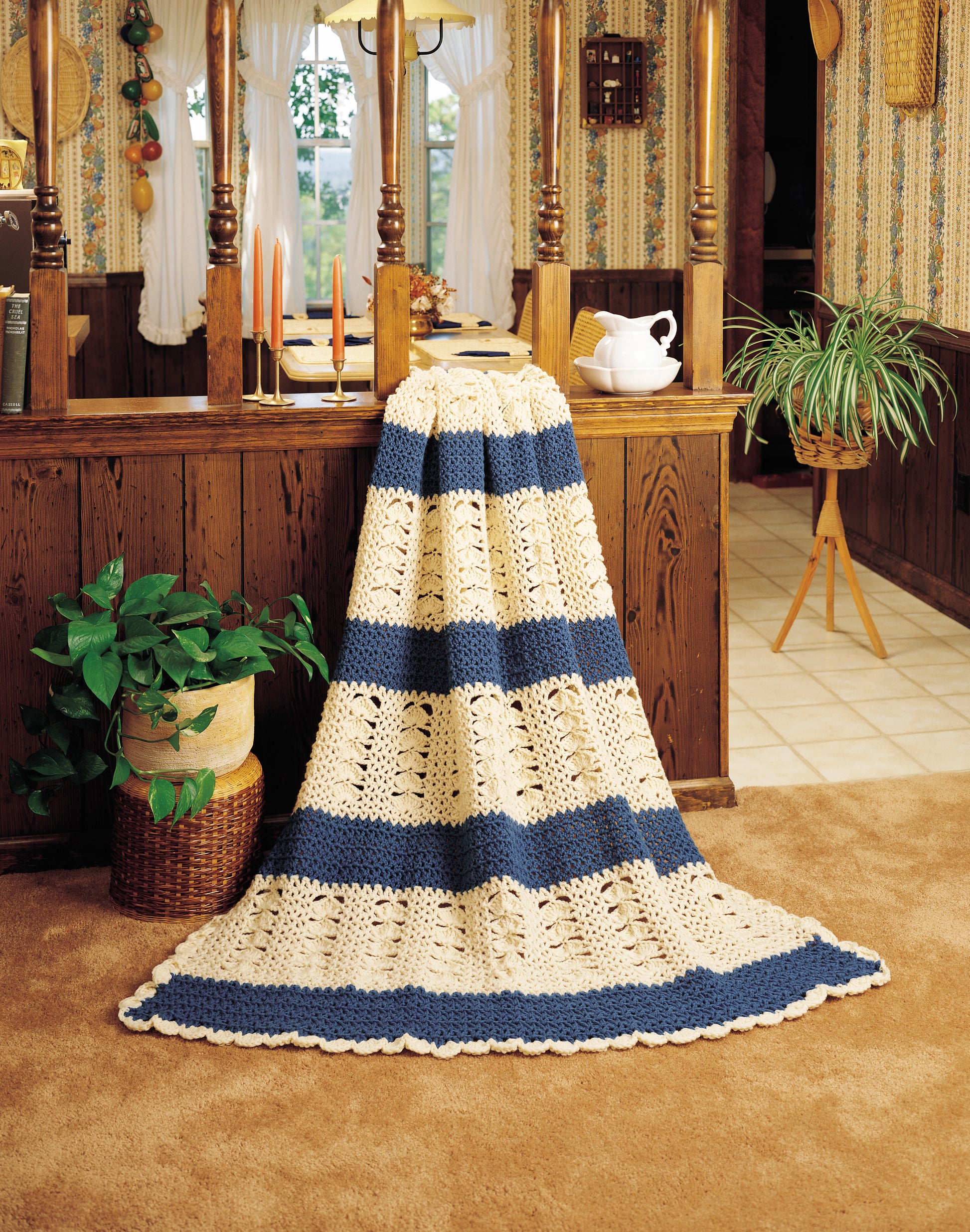 The Afghan Parade by Leisure Arts, a cream and blue striped worsted weight blanket, is draped over a wooden counter in a cozy vintage kitchen accented with plants, candles, and a teapot in the warm, carpeted space.