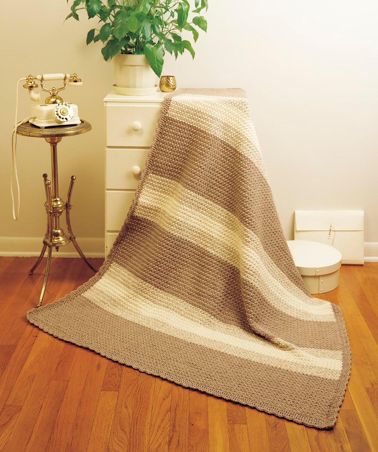 The Afghan Parade by Leisure Arts, a beige and cream striped knitted blanket for beginner crochet projects, is draped over a white dresser near a vintage rotary phone, brass table, green plant, and white box on a wooden floor.