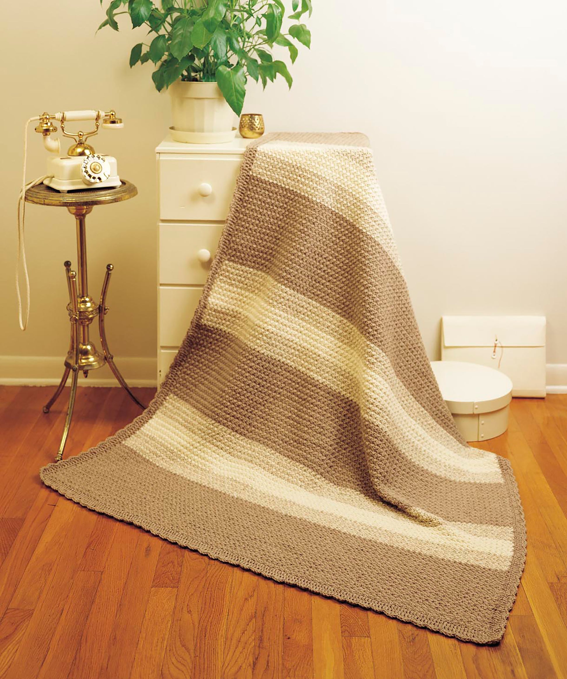 The Afghan Parade by Leisure Arts, a beige and cream striped knitted blanket for beginner crochet projects, is draped over a white dresser near a vintage rotary phone, brass table, green plant, and white box on a wooden floor.