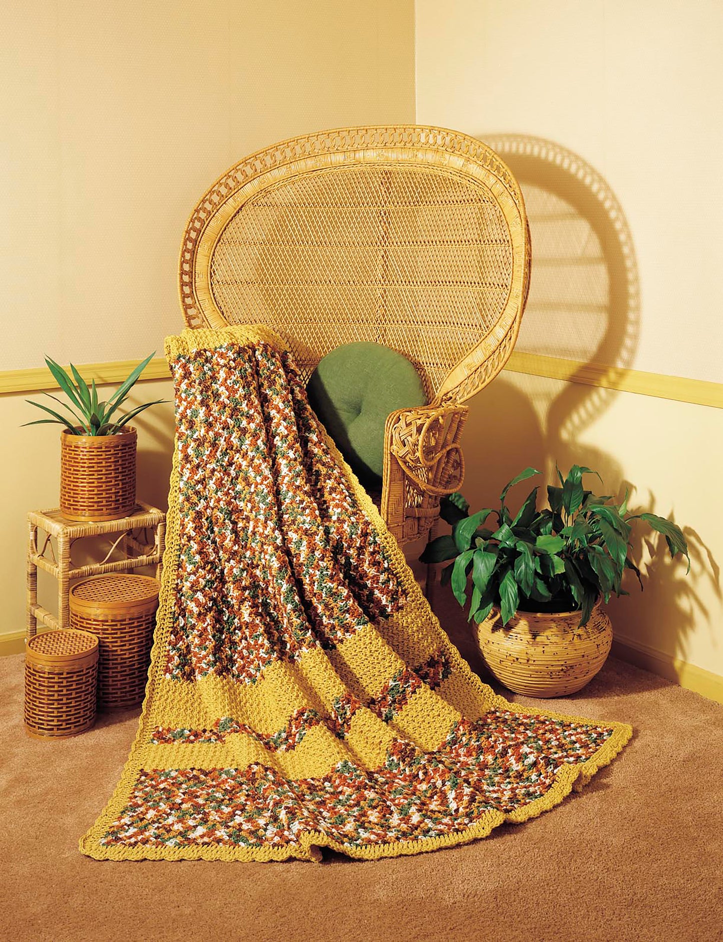 A wicker chair with a green cushion holds the Afghan Parade blanket by Leisure Arts, crafted from beginner crochet projects. Next to it are stacked wicker boxes topped with a potted plant and another green plant in a patterned ceramic pot.