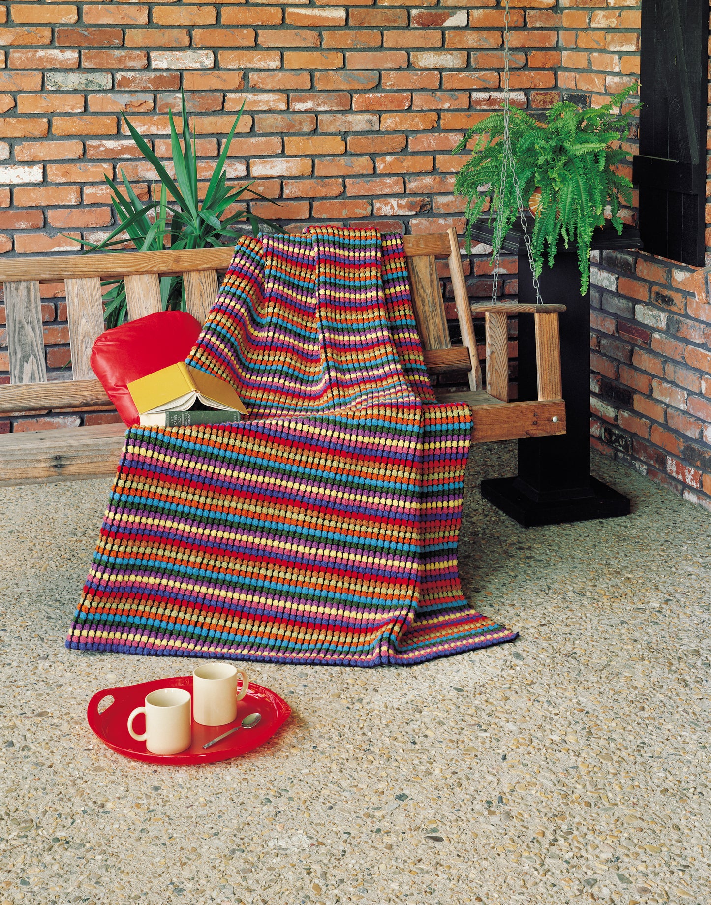 The Afghan Parade crochet blanket by Leisure Arts adds color to a wooden bench with a red pillow and books on a stone floor. A red tray with two mugs and a teapot sits nearby, surrounded by green plants and a brick wall for a cozy touch.