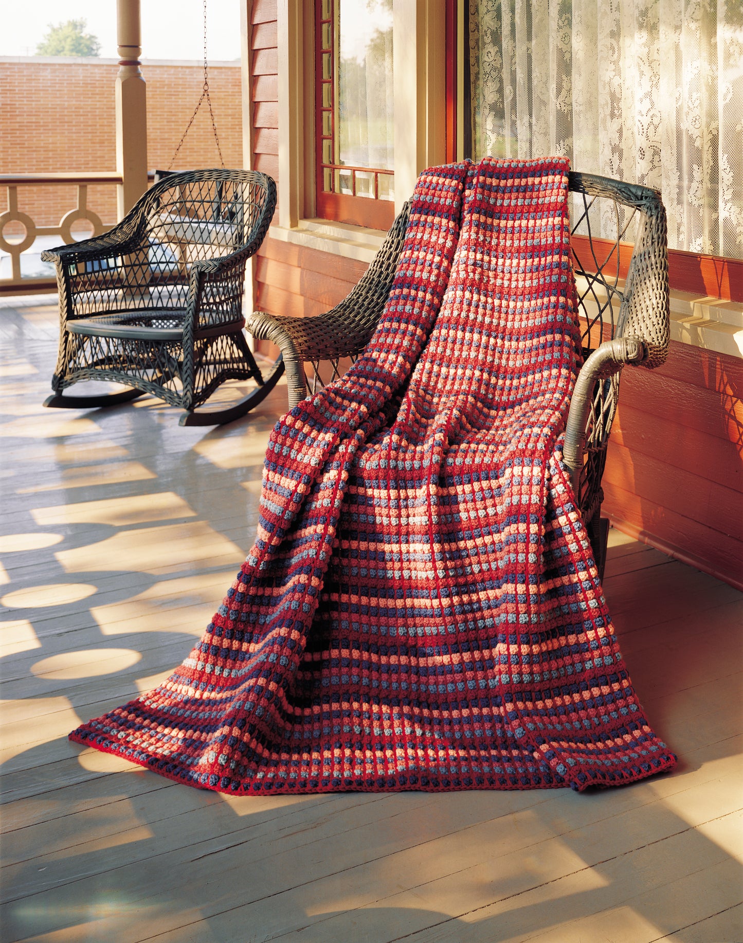 The Afghan Parade by Leisure Arts, a handmade striped blanket in red and purple worsted weight yarn, is draped over a wicker rocking chair on a sunlit porch, with another empty rocker nearby and sunlight streaming in.