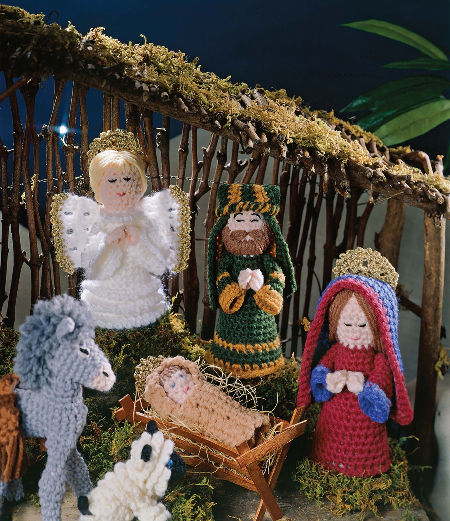 Leisure Arts’ "Away in a Manger—A Crochet Nativity" set features handmade worsted yarn figures—Mary, Joseph, baby Jesus in a manger, an angel, shepherd, donkey, and sheep—in a stable adorned with moss and wooden sticks.