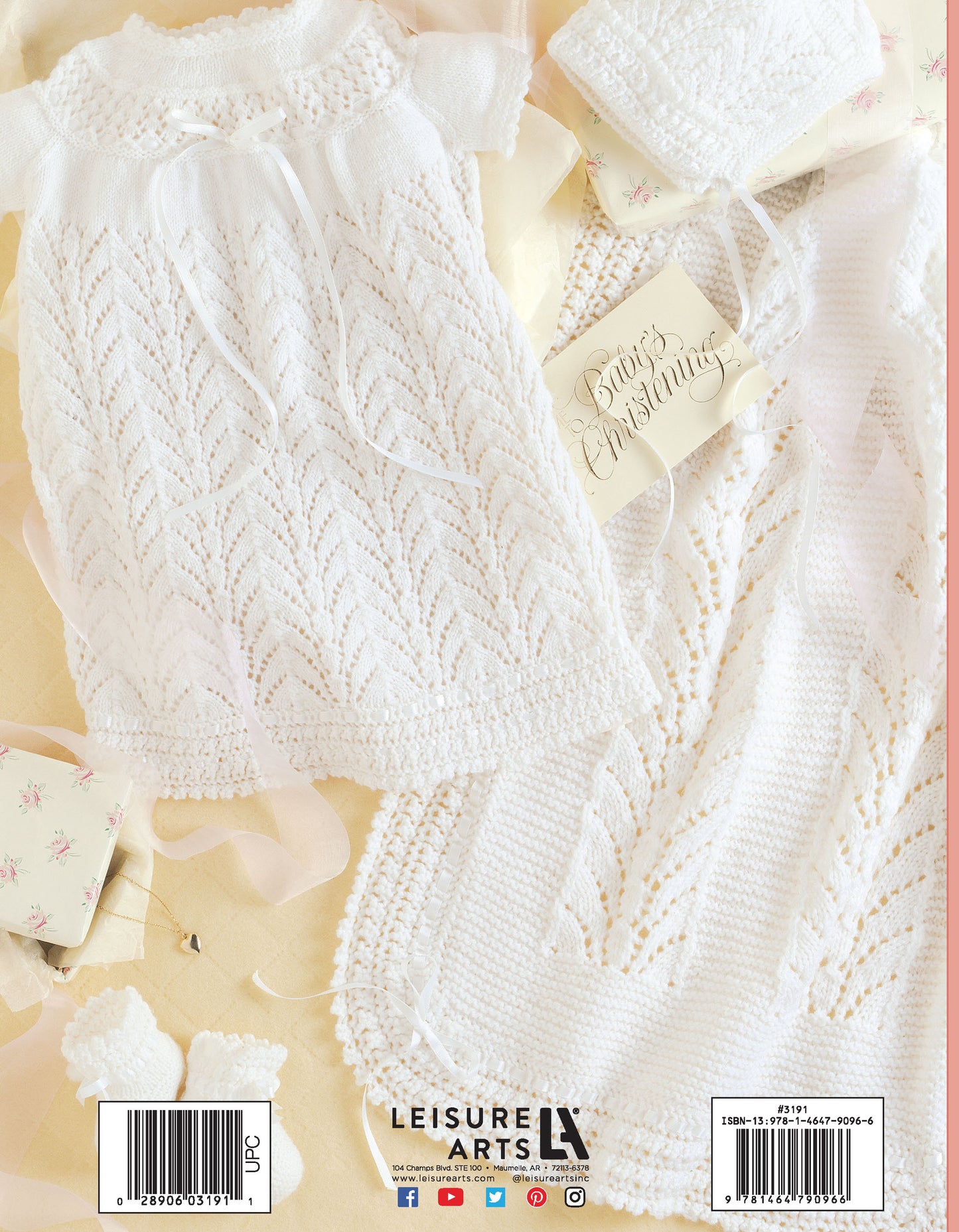 A delicate white Knit Layette by Leisure Arts, featuring a dress, bonnet, and blanket crafted from superfine yarn, is arranged on a pale surface with a “Baby’s Christening” tag and floral gift boxes nearby.