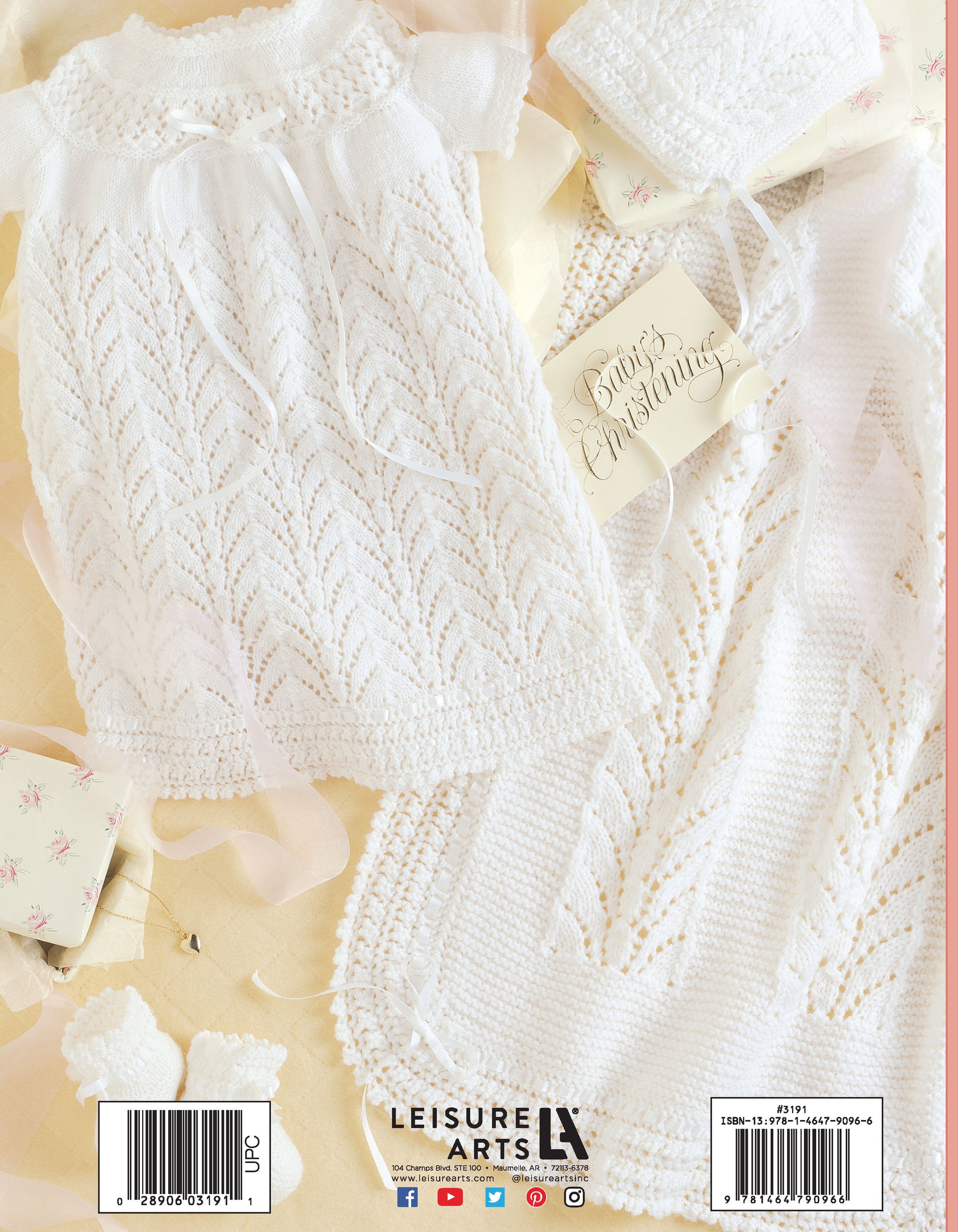 A delicate white Knit Layette by Leisure Arts, featuring a dress, bonnet, and blanket crafted from superfine yarn, is arranged on a pale surface with a “Baby’s Christening” tag and floral gift boxes nearby.