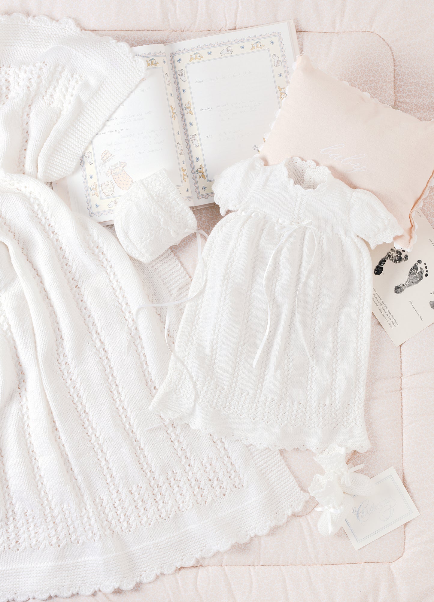 The Knit Layette by Leisure Arts features white baby items—dress, bonnet, blanket, and shoes—arranged on a pink quilt with a memory book, footprint card, and pillow for a charming newborn set display.
