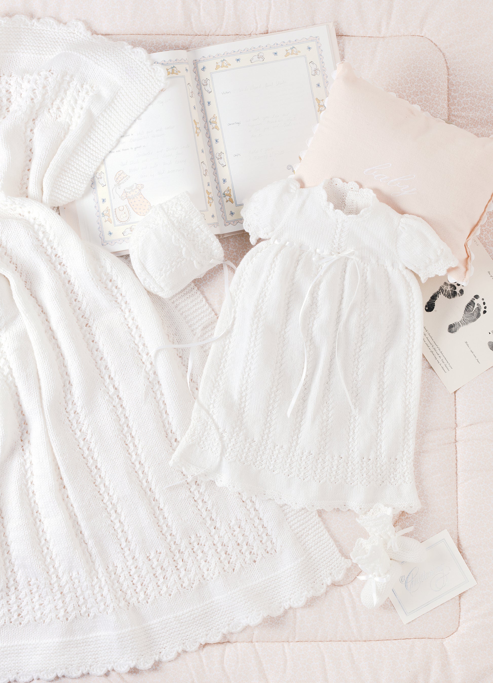 The Knit Layette by Leisure Arts features white baby items—dress, bonnet, blanket, and shoes—arranged on a pink quilt with a memory book, footprint card, and pillow for a charming newborn set display.