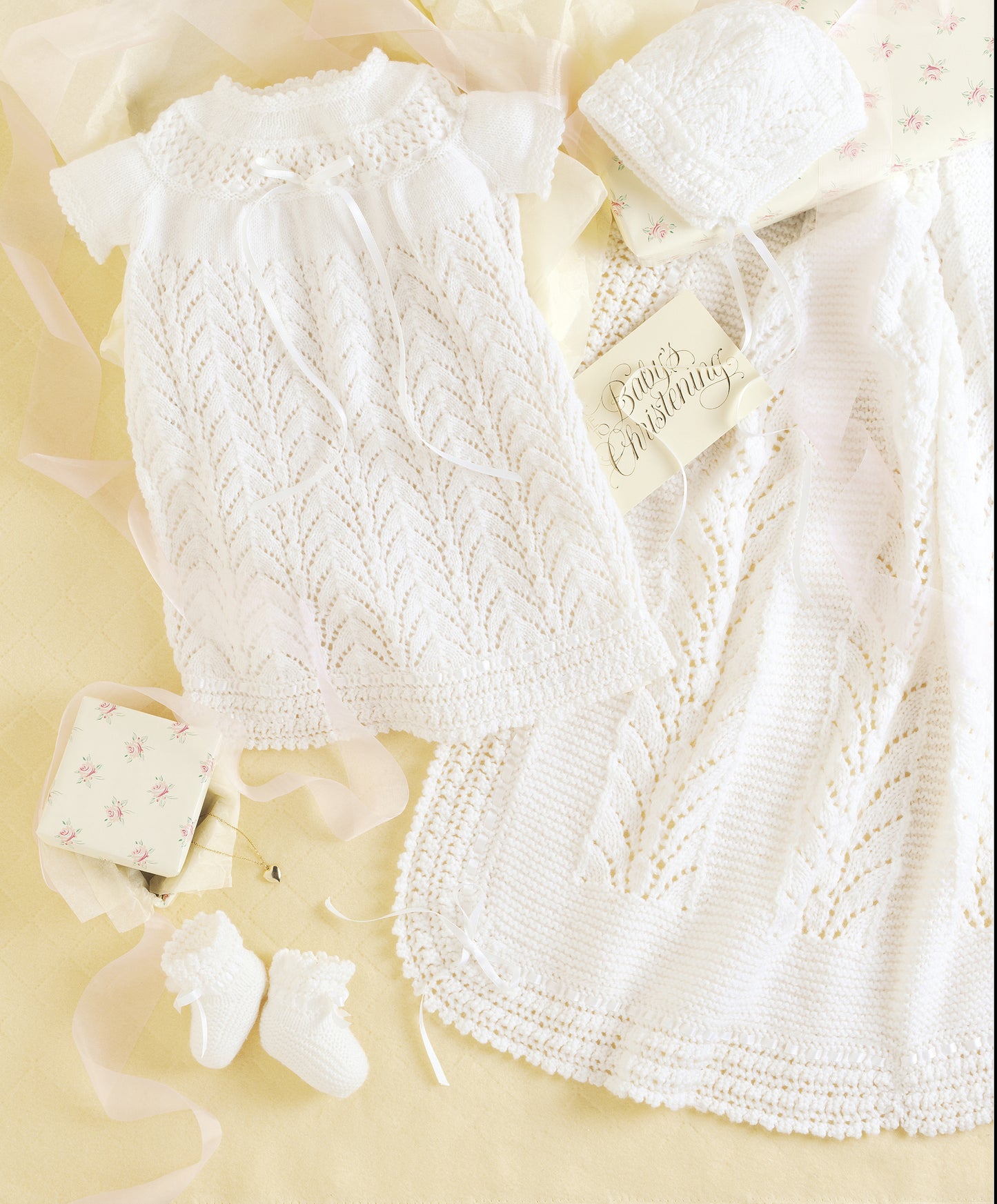 The Leisure Arts Knit Layette includes a delicate white newborn gown, bonnet, booties, and blanket made with superfine yarn, shown on a pale yellow background with ribbons and a "Baby's Christening" card.