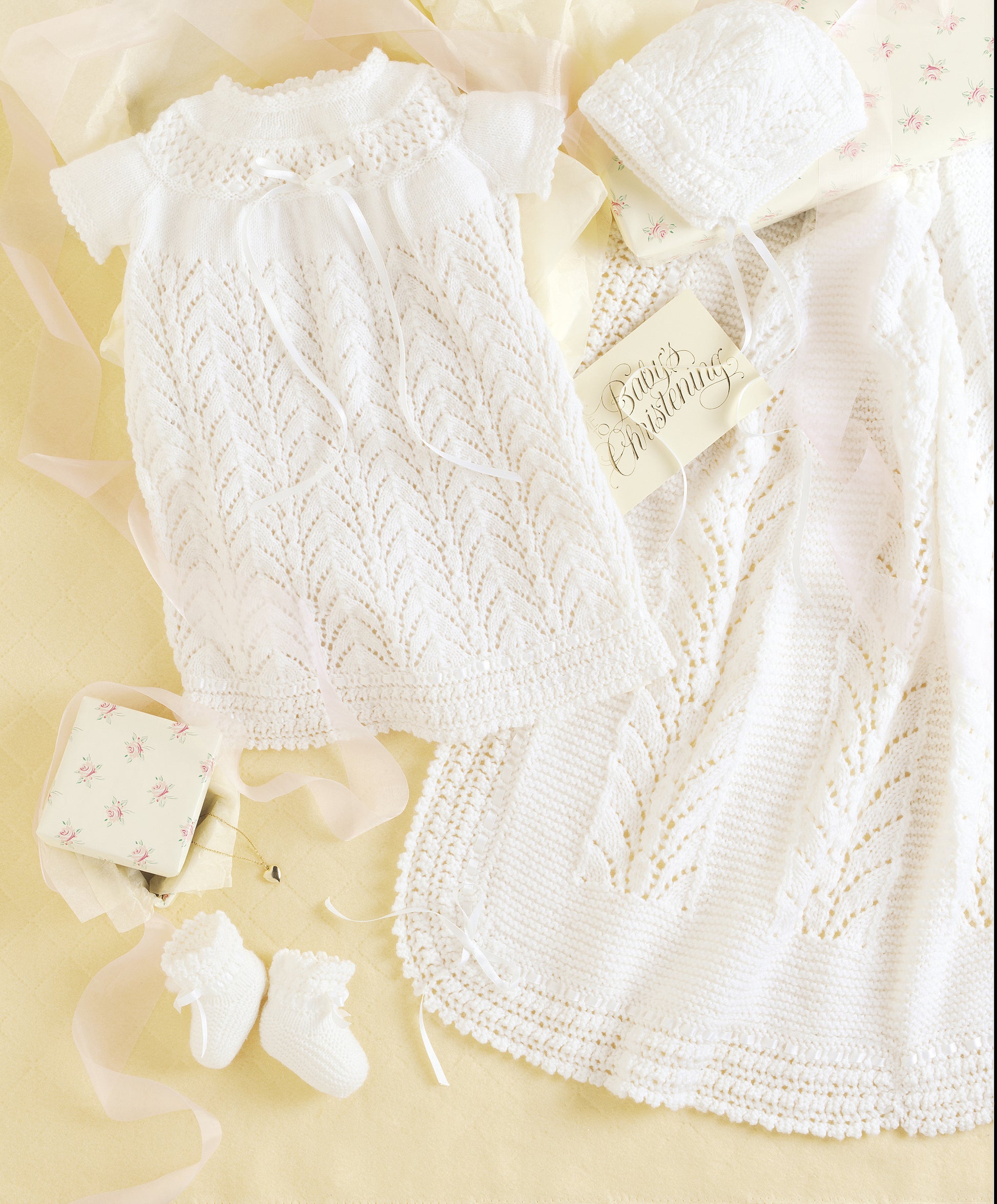 The Leisure Arts Knit Layette includes a delicate white newborn gown, bonnet, booties, and blanket made with superfine yarn, shown on a pale yellow background with ribbons and a "Baby's Christening" card.