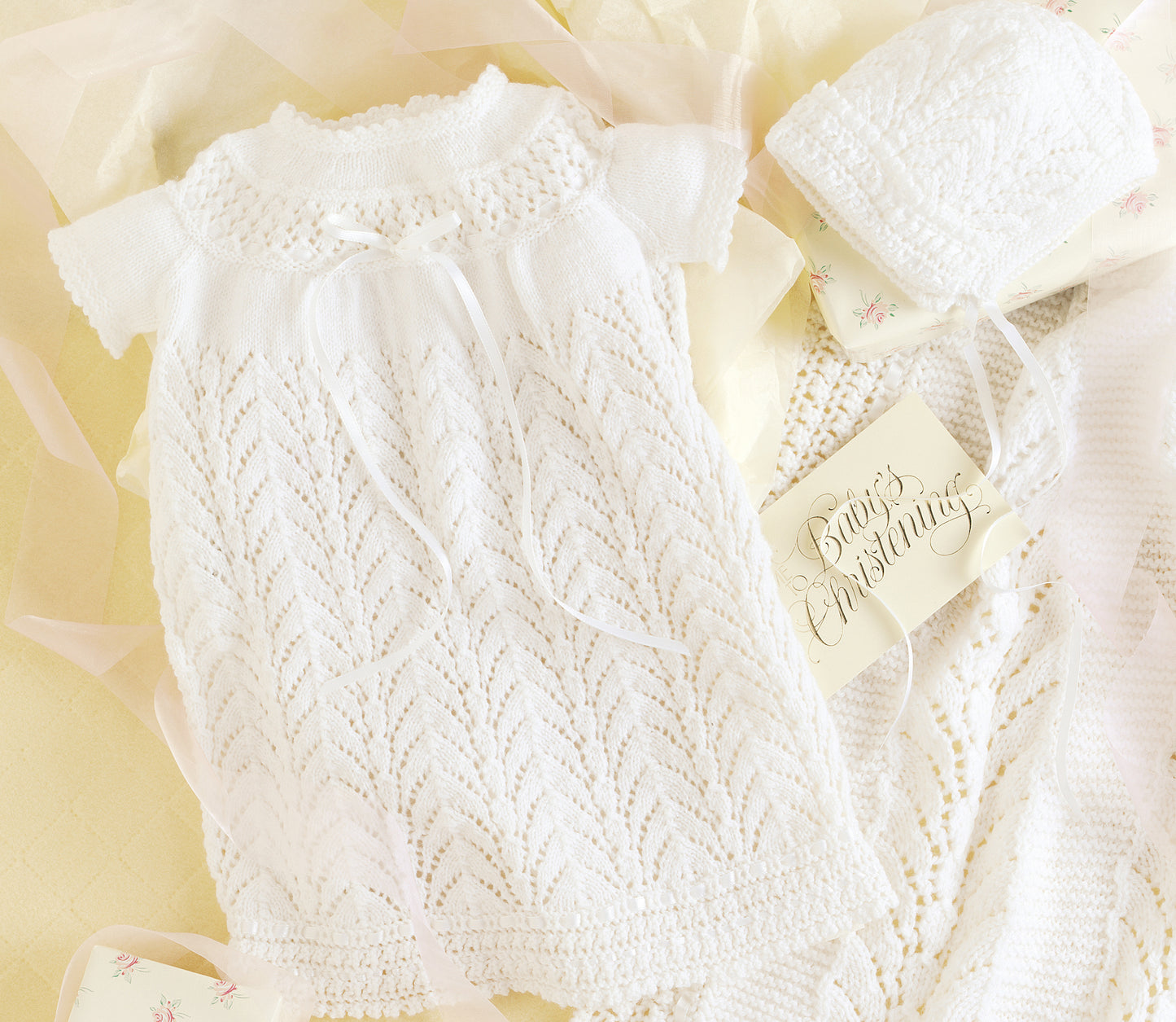 The Leisure Arts Knit Layette is a delicate white newborn gown set with a matching bonnet in intricate lace, displayed on soft yellow fabric beside a card reading "Baby's Christening.