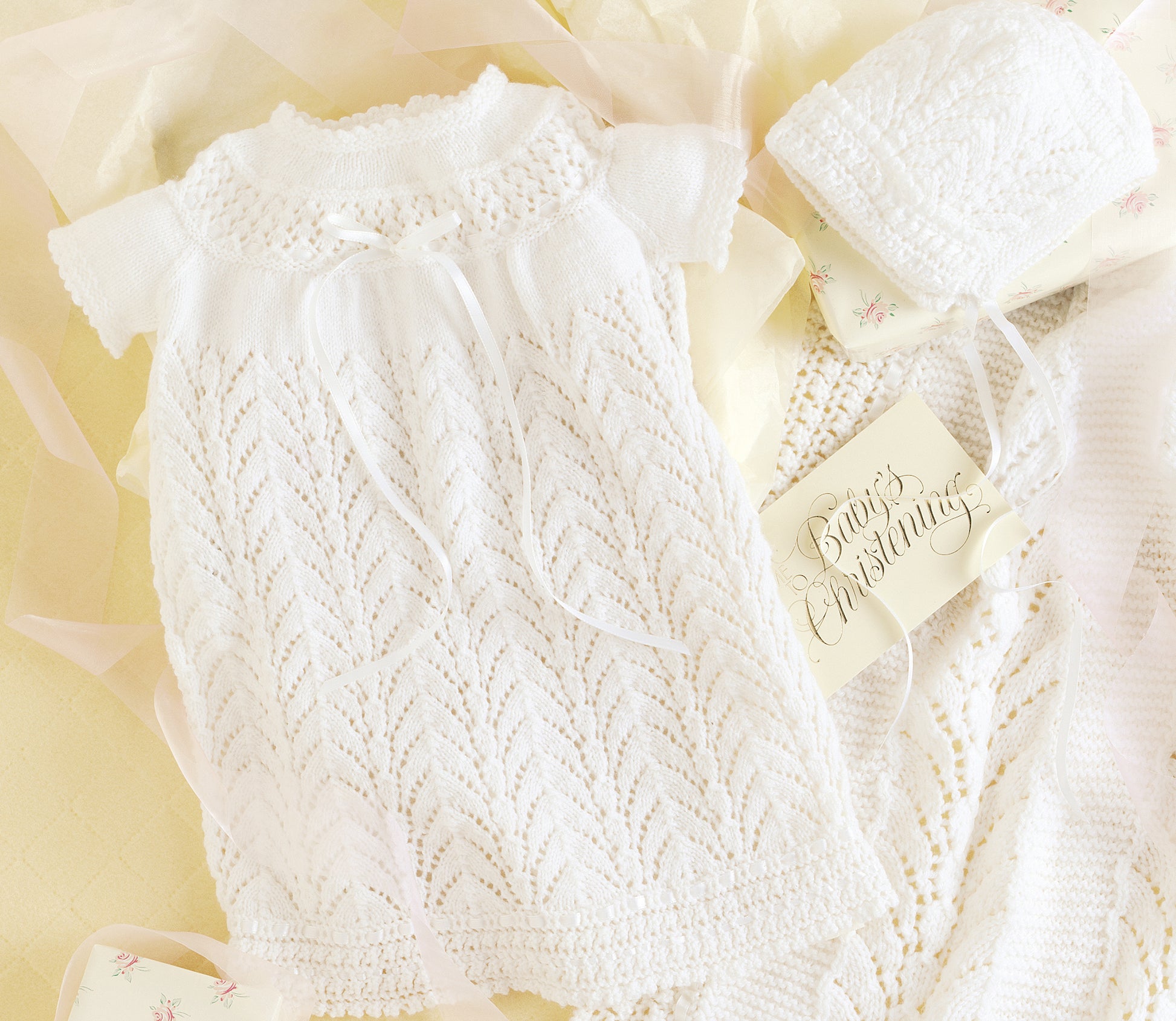 The Leisure Arts Knit Layette is a delicate white newborn gown set with a matching bonnet in intricate lace, displayed on soft yellow fabric beside a card reading "Baby's Christening.