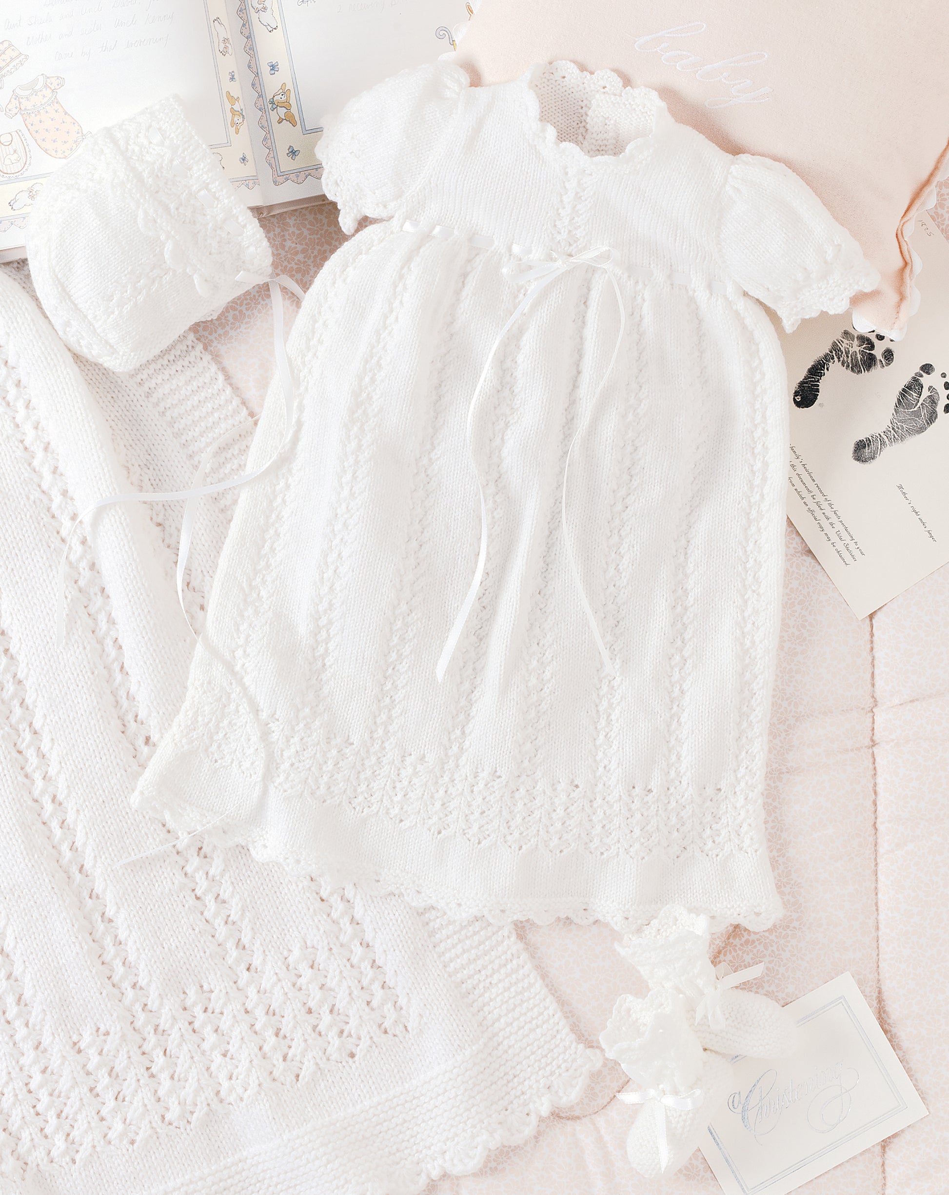 The Leisure Arts Knit Layette, featuring a delicate white knitted baby dress with short sleeves and ribbon ties, is displayed with a matching bonnet, blanket, and card among open baby books and a pink pillow on a pastel background.